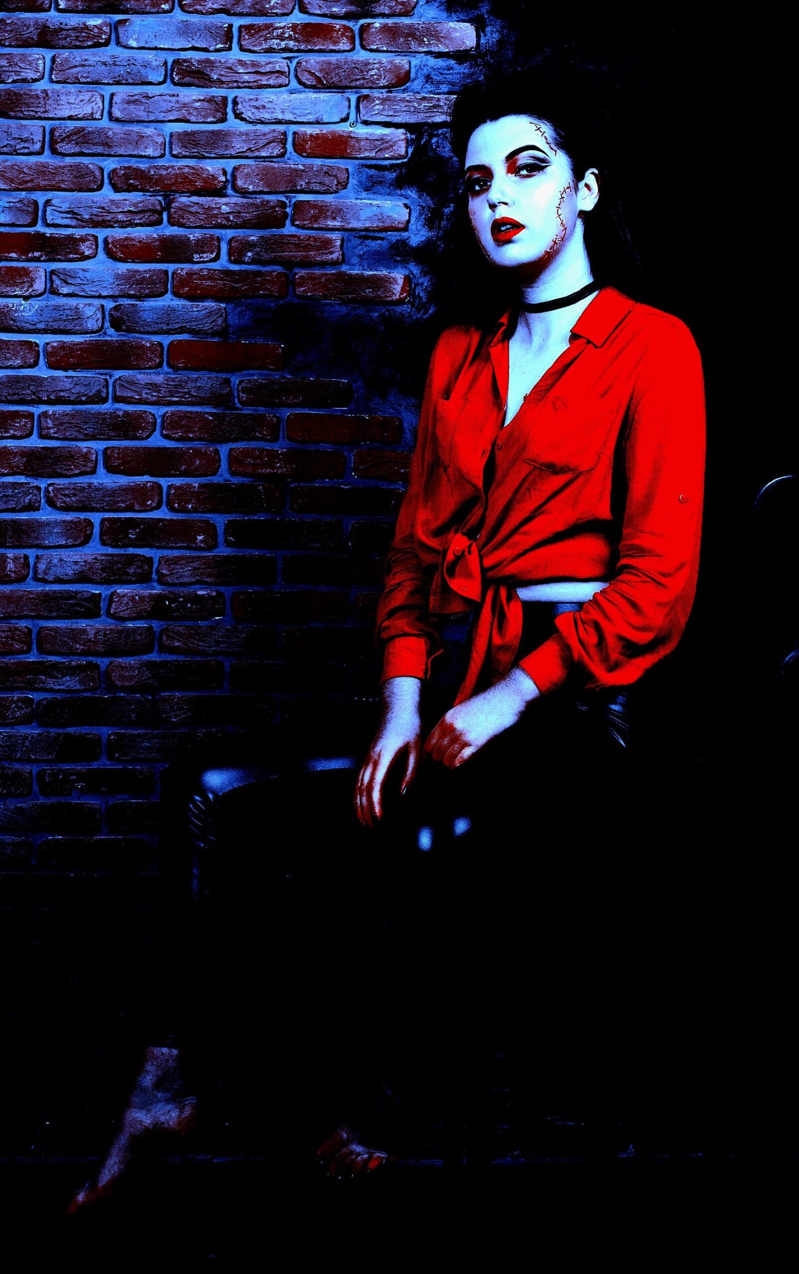 Gothic woman in a dramatic pose against a dark background, wearing a red button-down shirt and black leather pants, exuding a seductive and mysterious aura with bold makeup.