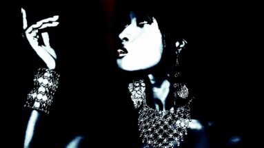 Gothic woman standing by a ladder, wearing dark makeup and black clothing, set against a vibrant blue background.