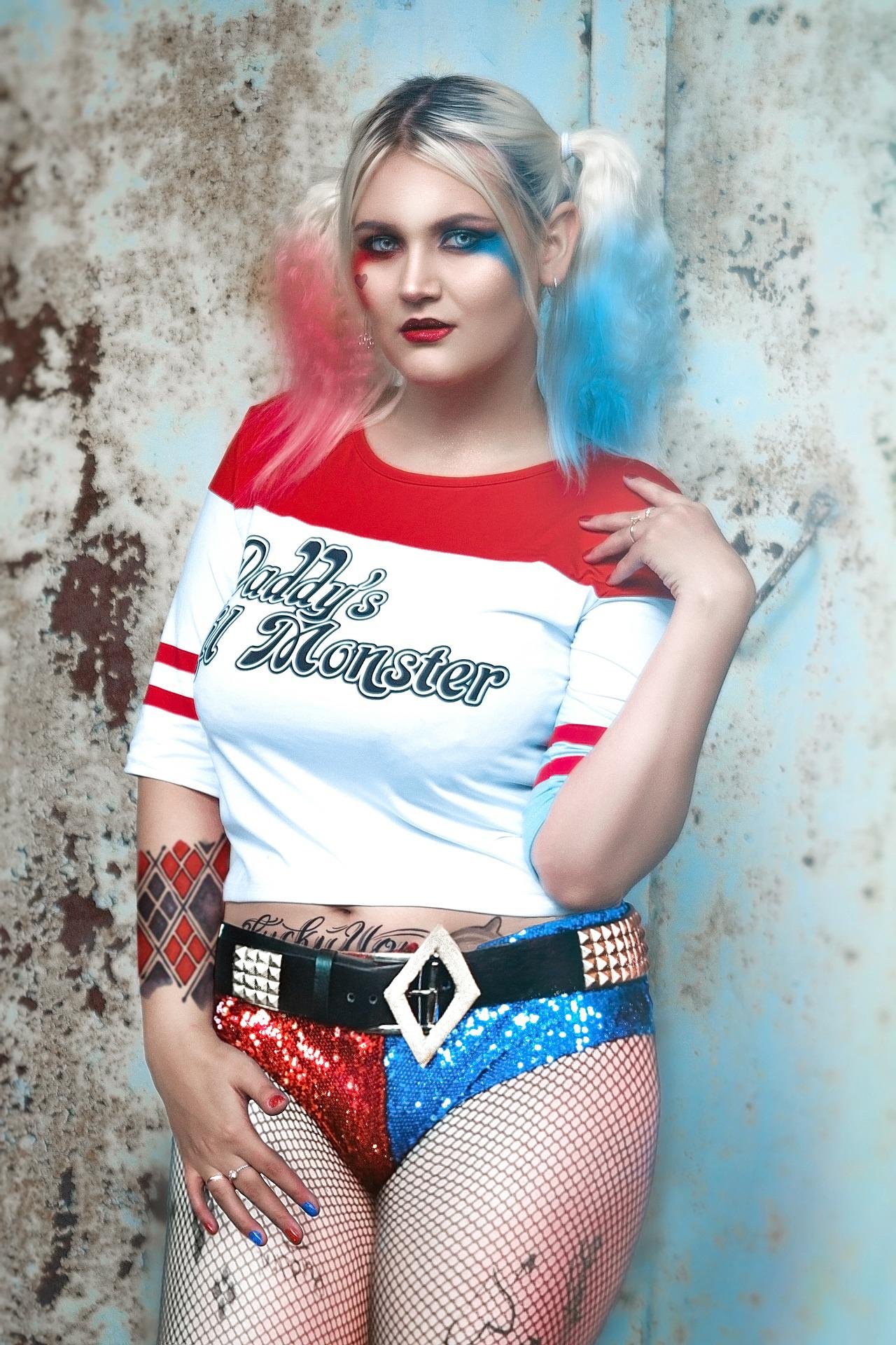Woman cosplaying as Harley Quinn in a playful pose, wearing the iconic "Daddy's Lil Monster" shirt with blue and pink pigtails.