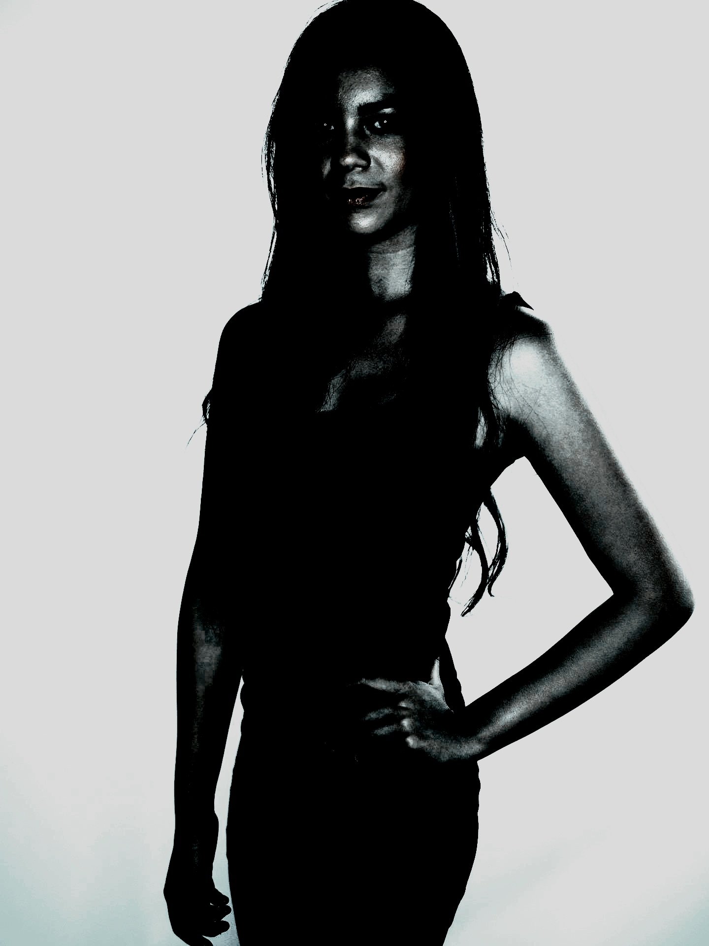 A dark, high-contrast portrait of a woman standing confidently with one hand on her hip, exuding a strong and shadowy presence.
