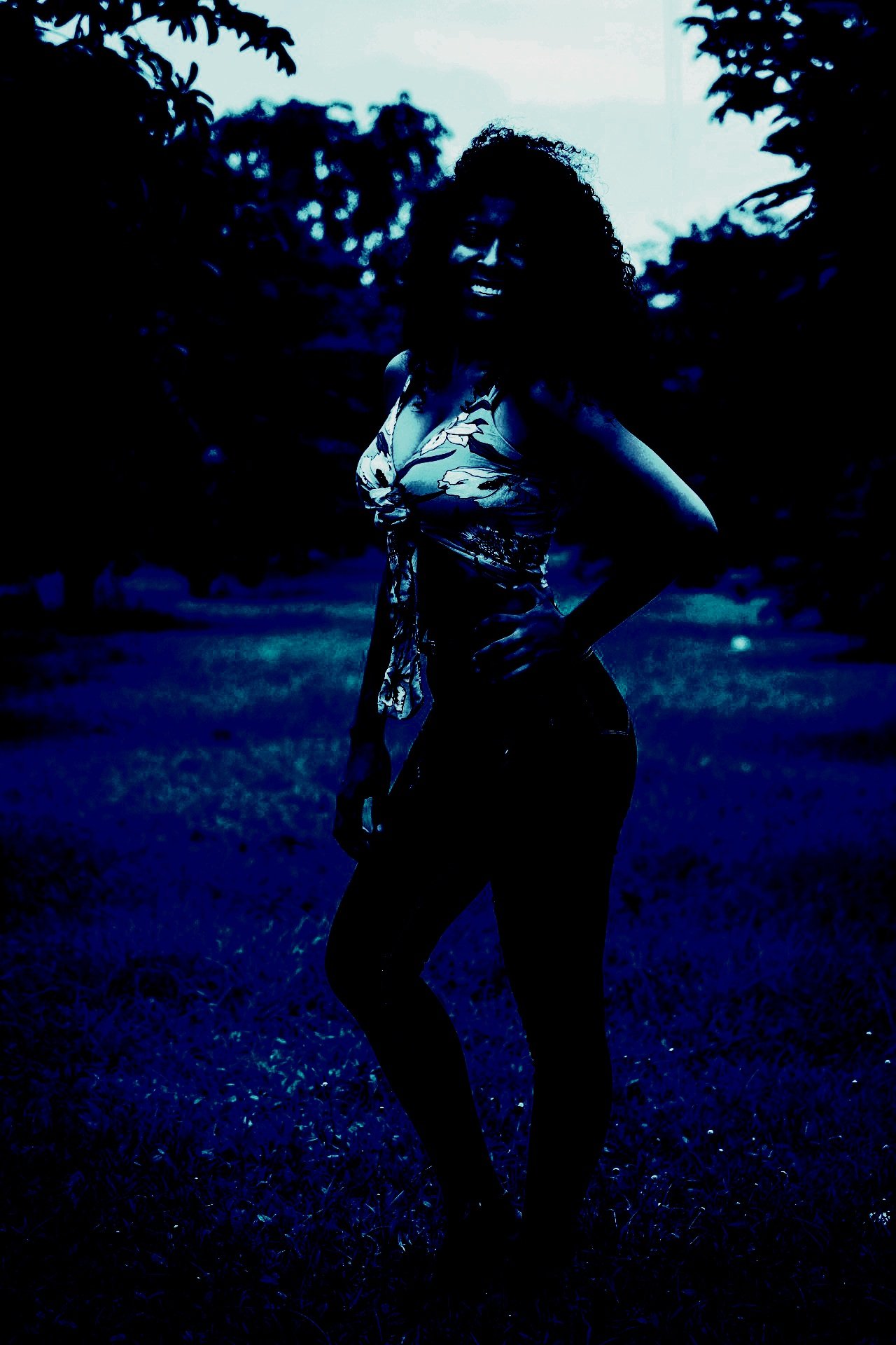 A woman standing outdoors in a dimly lit setting, with a dramatic shadowed effect, conveying a mysterious and intense mood.