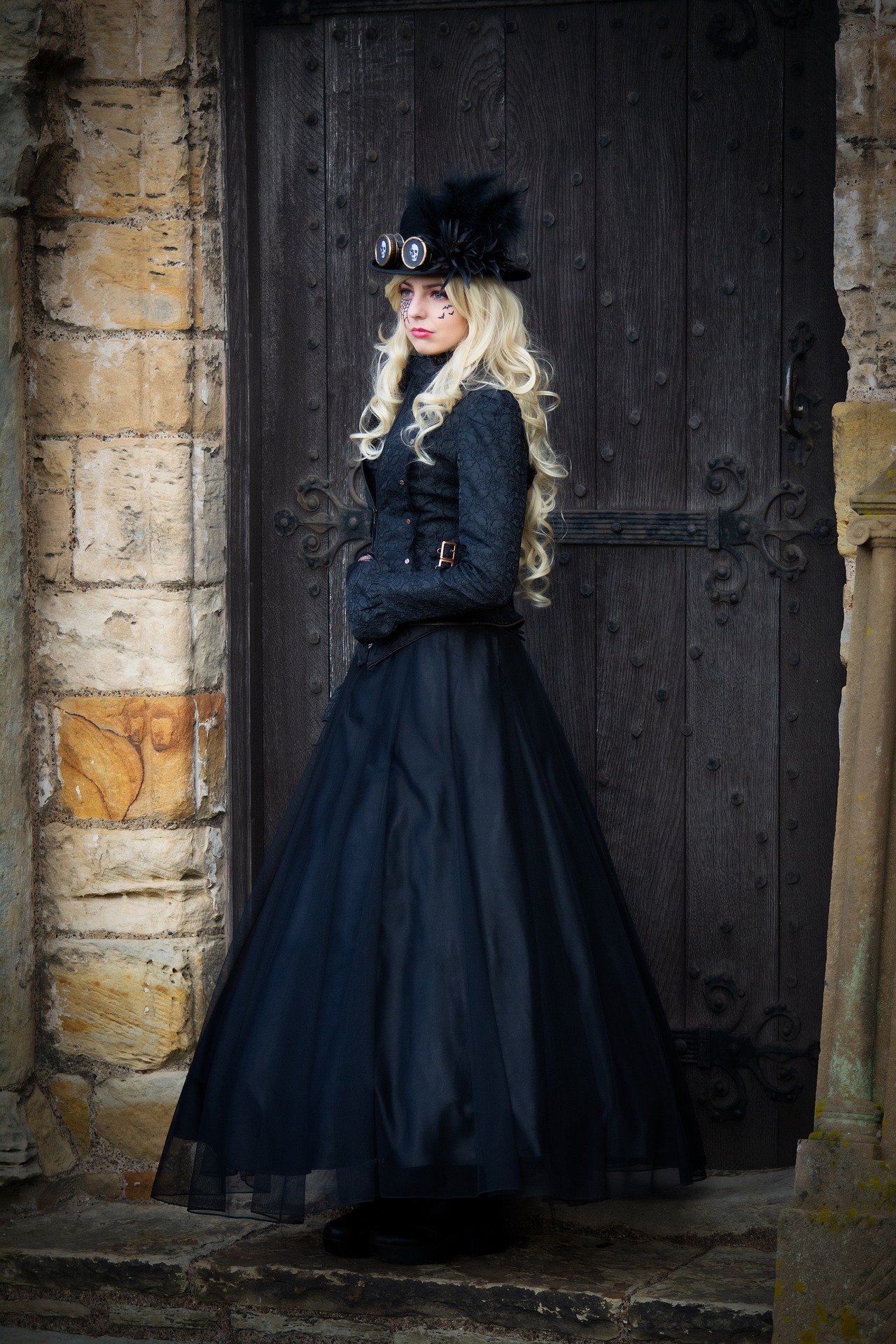 Blonde woman in a long, black gothic steampunk gown with feathered hat and goggles, standing in front of an old wooden door.