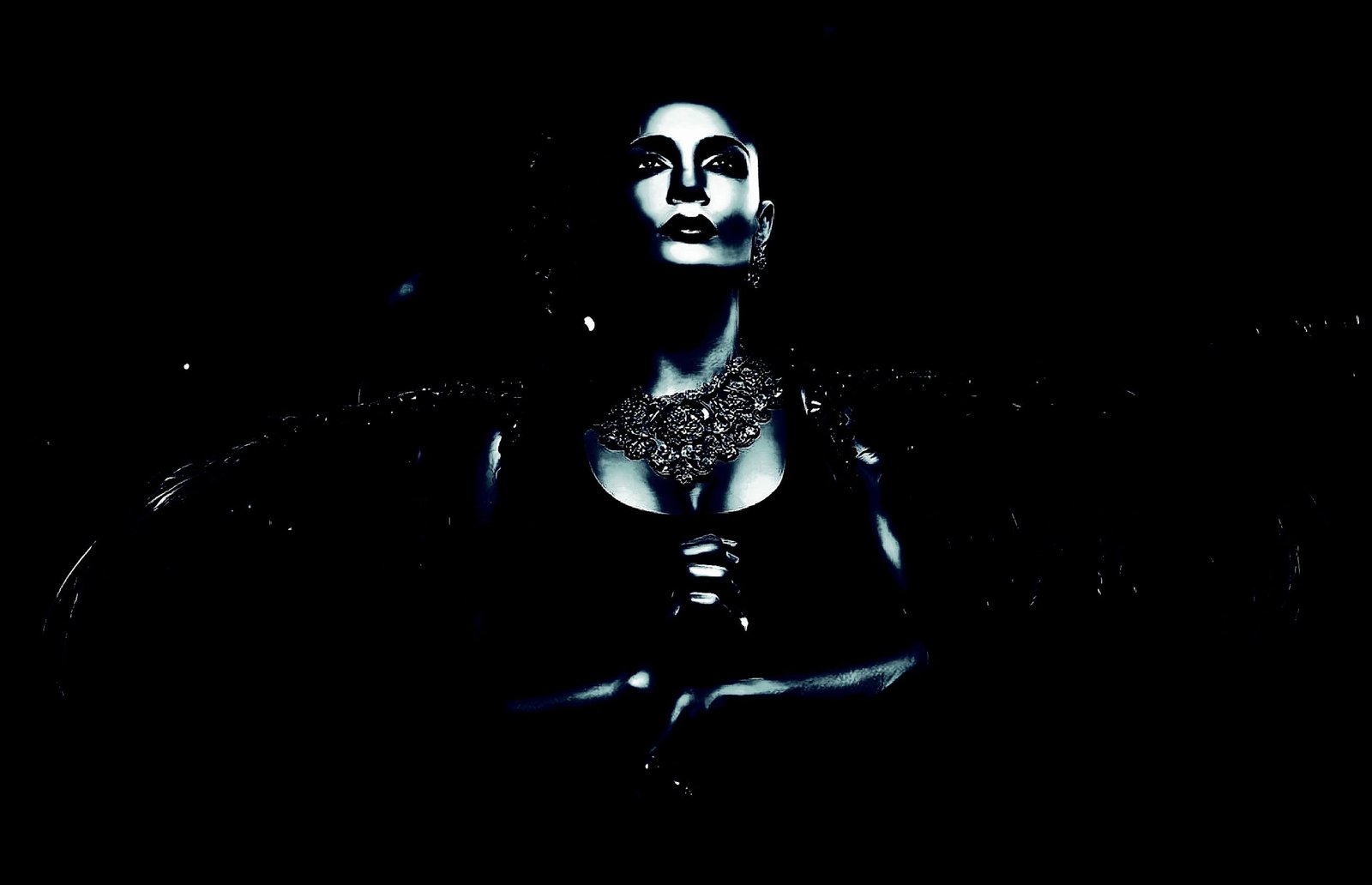 Woman with dramatic makeup, dressed in dark attire, standing with a powerful presence against a pitch-black background.