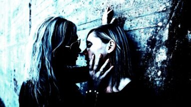 Two women in a close, intimate moment against a textured wall, bathed in a blue-toned light.