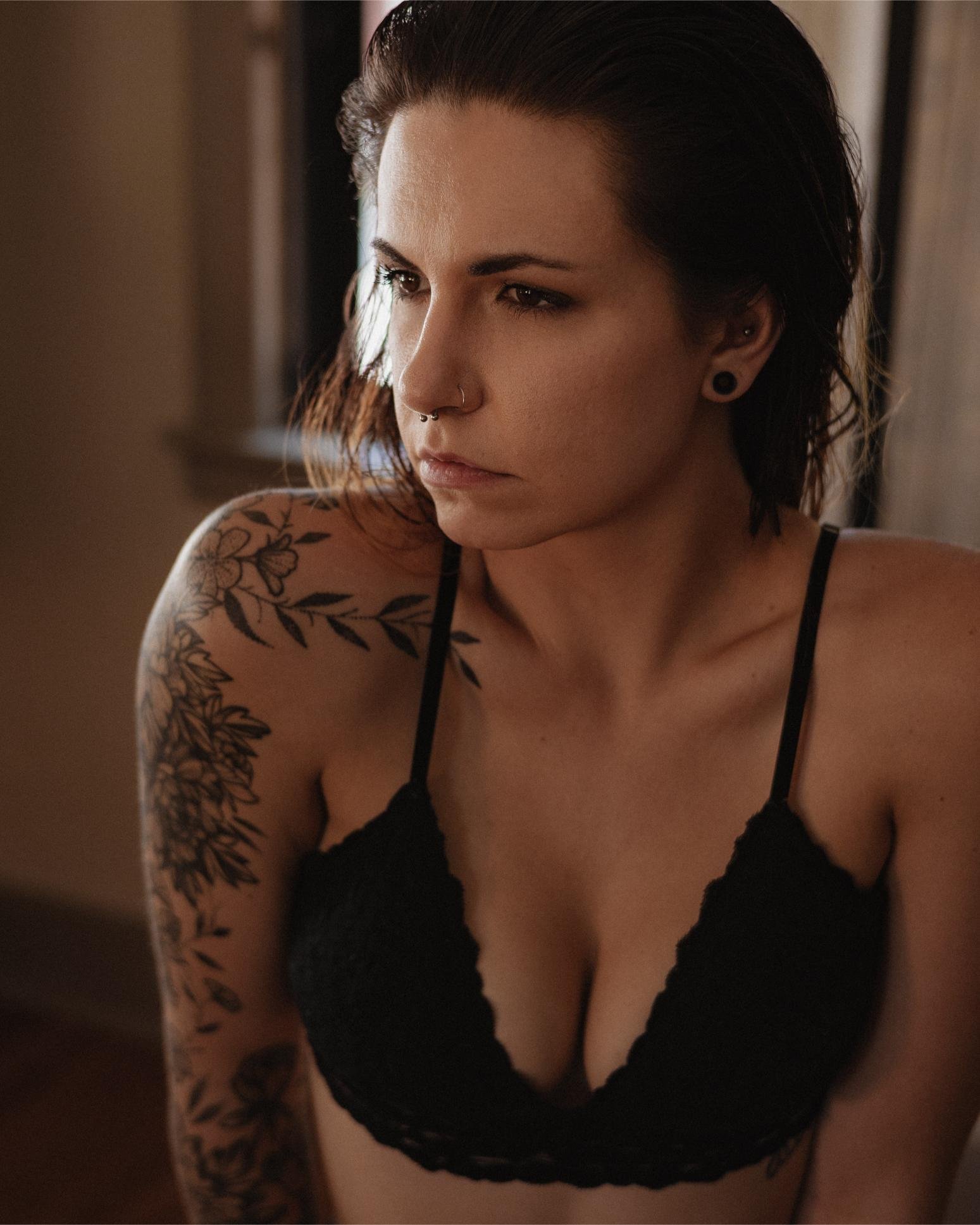 Serious-looking woman with tattoos on her arm, wearing a black bralette, sitting by a window with soft natural light.