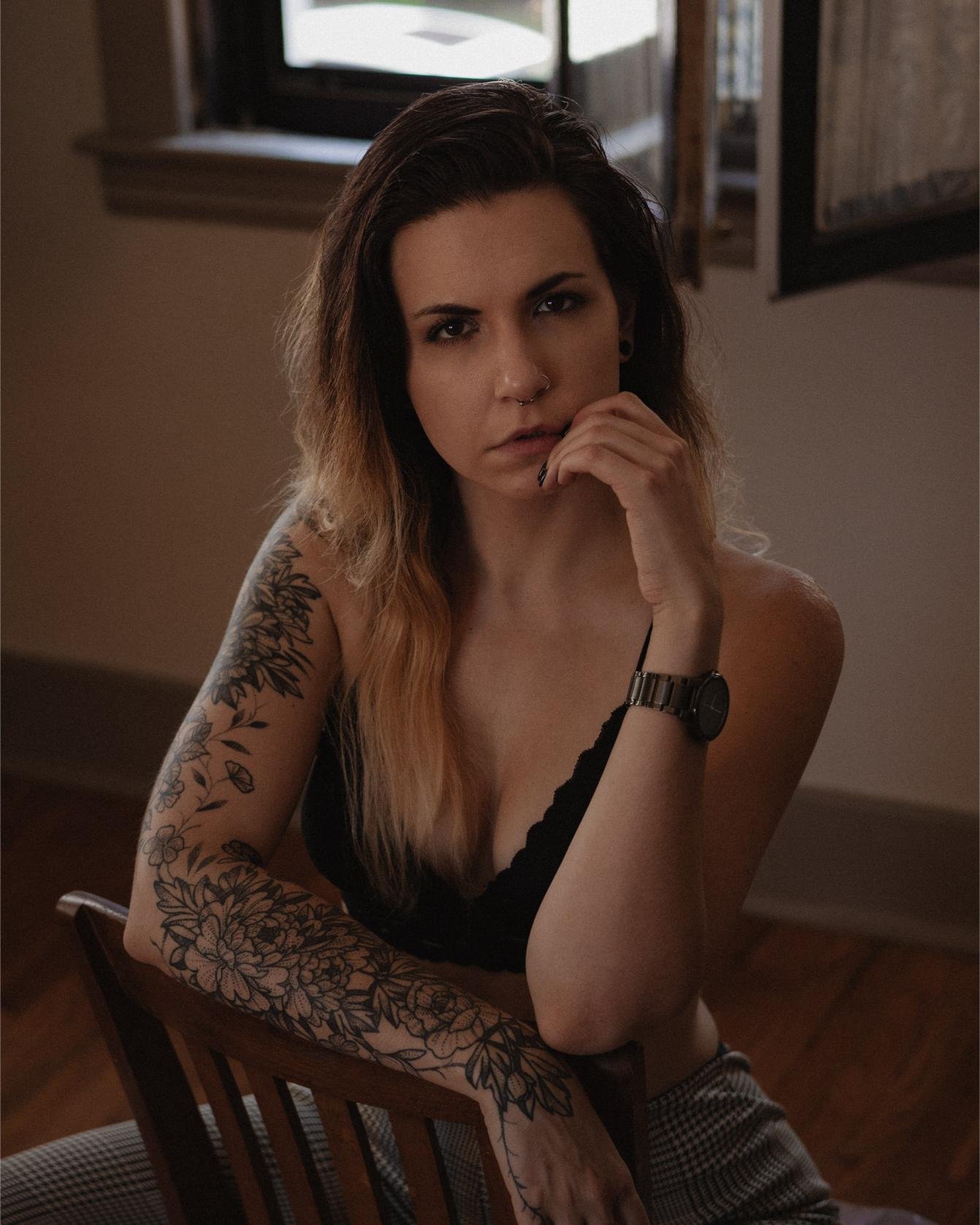 Pensive woman with long, wavy hair, resting her chin on her hand, showcasing her arm tattoos and a minimalistic black outfit.