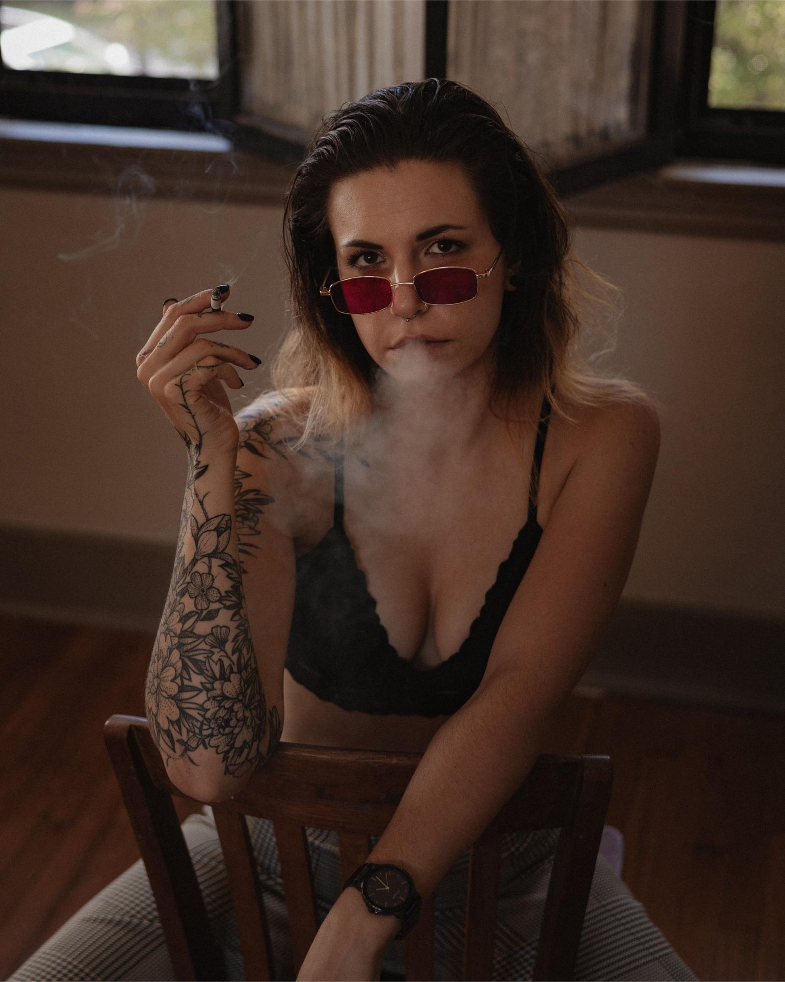 Woman with red sunglasses, sitting on a wooden chair, smoking, with tattoos on her arm and a confident gaze in a dimly lit room.