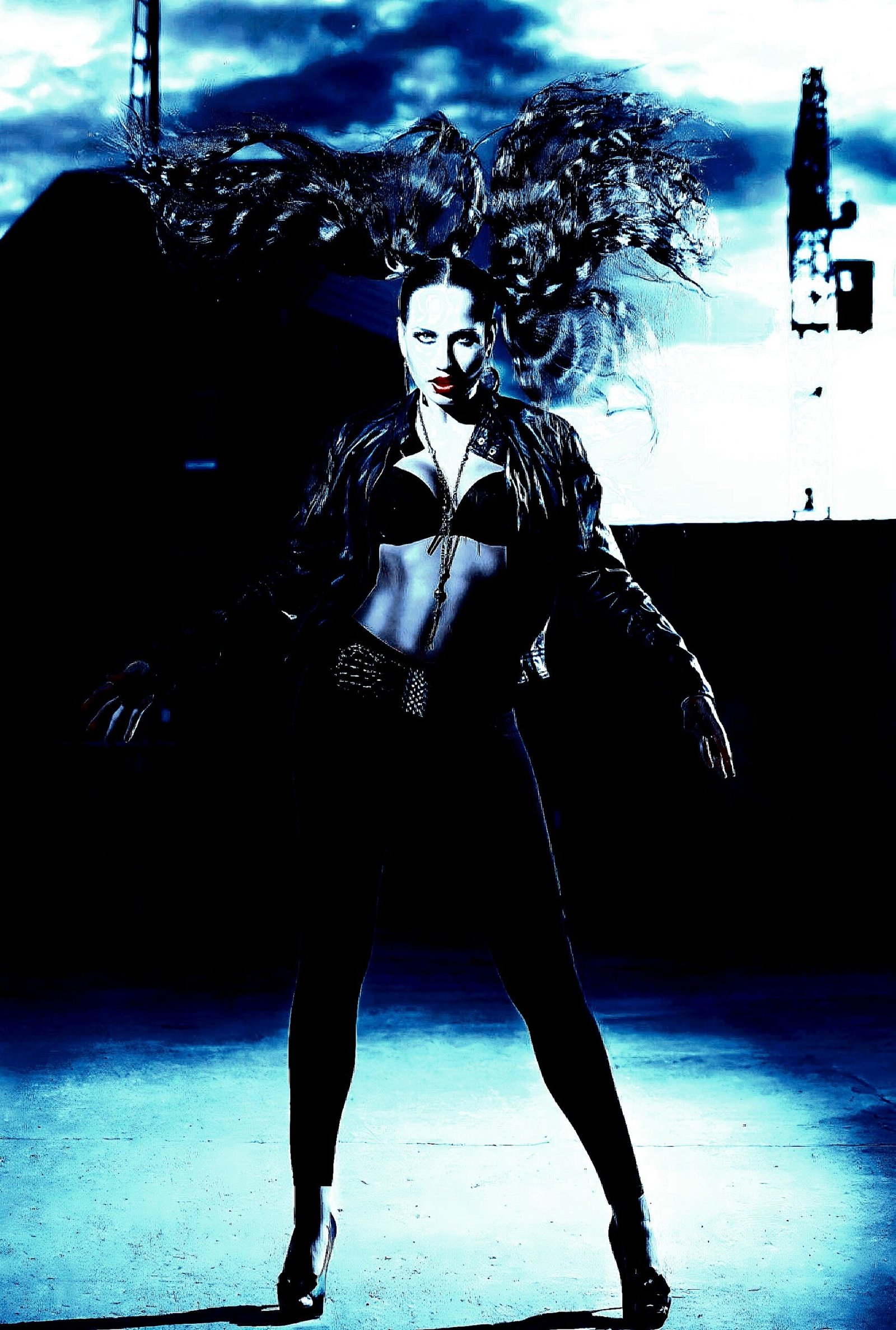 Woman in a black outfit with wild, flowing hair, standing confidently in an industrial outdoor setting.
