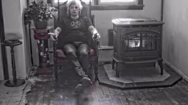 Man wearing a pentagram shirt, sitting in an ornate chair next to a vintage fireplace, in a shadowy room.