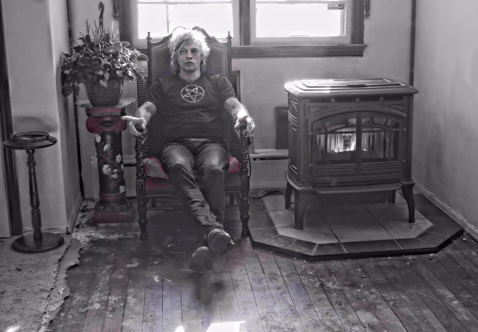 Man wearing a pentagram shirt, sitting in an ornate chair next to a vintage fireplace, in a shadowy room.