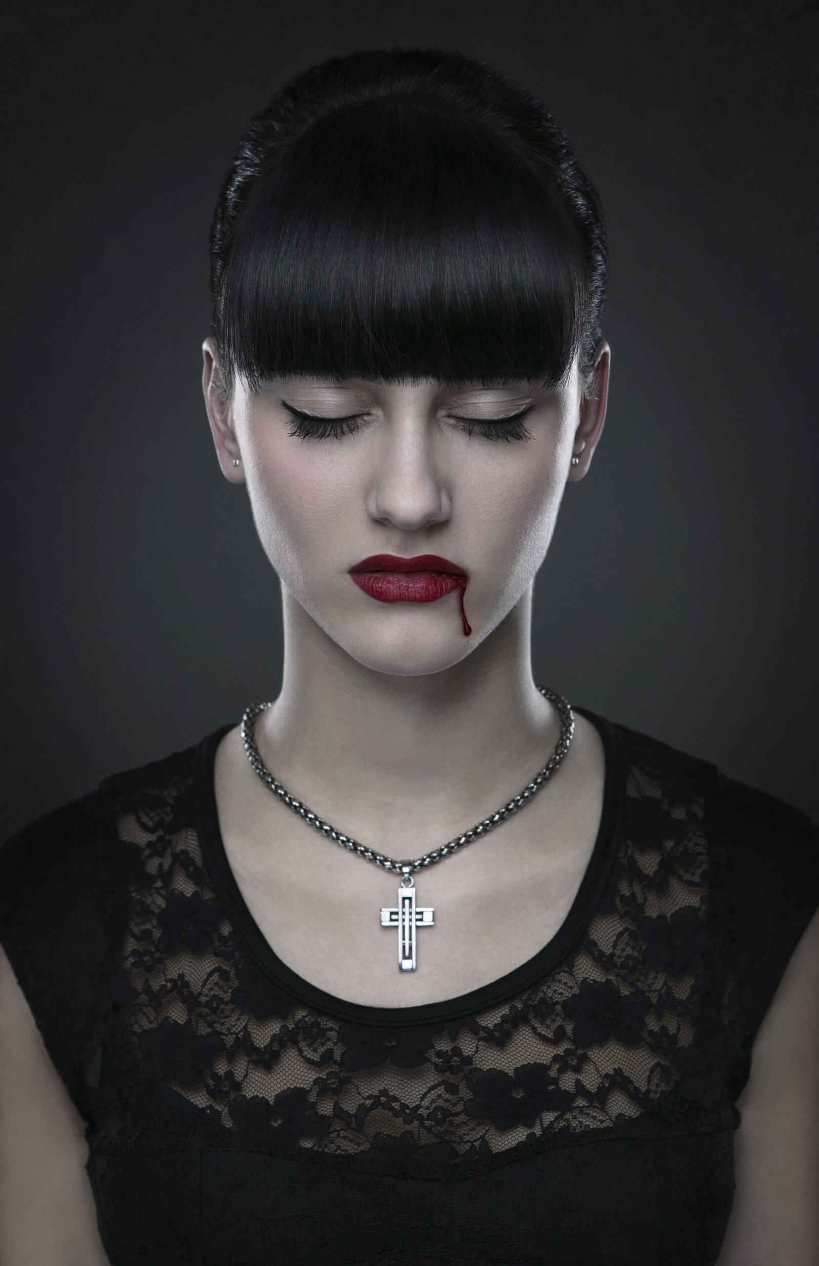 Woman with black hair and red lips, wearing a cross necklace, with a small drop of blood on her lip.