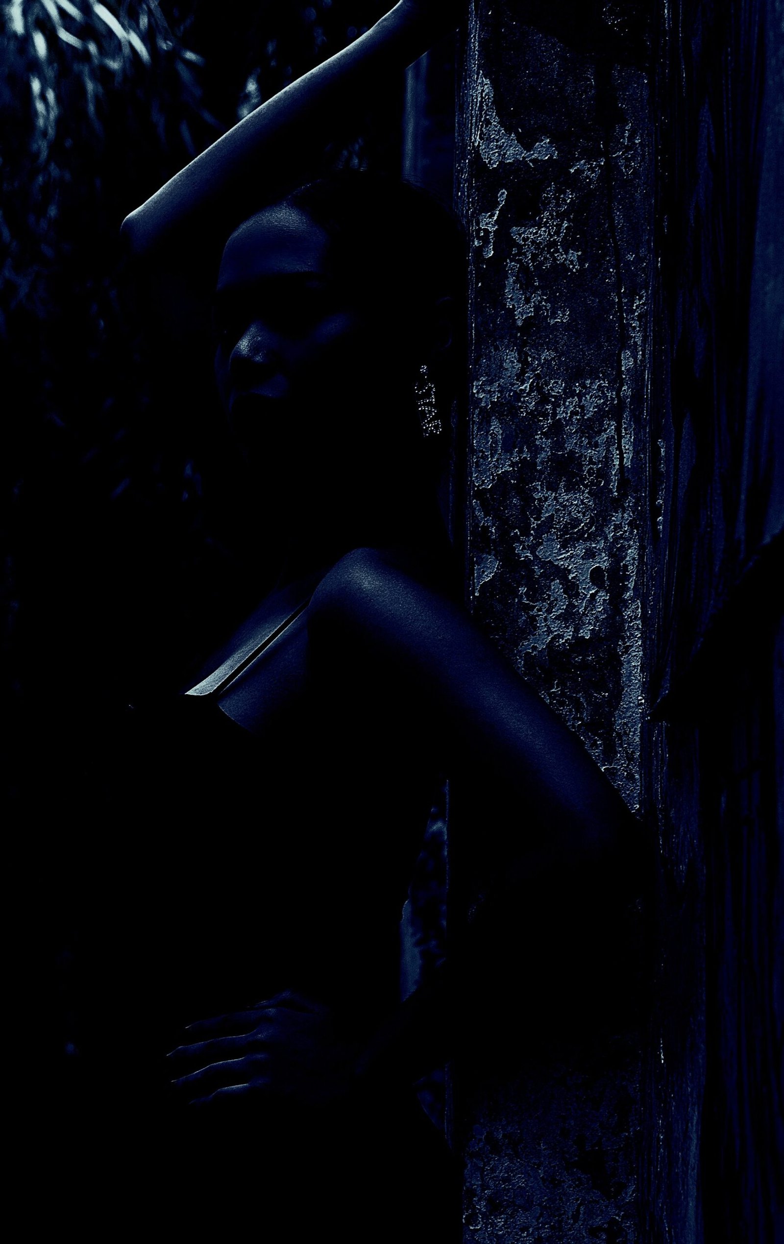 A mysterious silhouette of a woman leaning against a textured wall in a shadowy outdoor setting.
