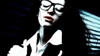 Close-up of a woman wearing oversized glasses, with bold red lips and her eyes closed, exuding confidence and sensuality, set against a dramatic, shadowy background.