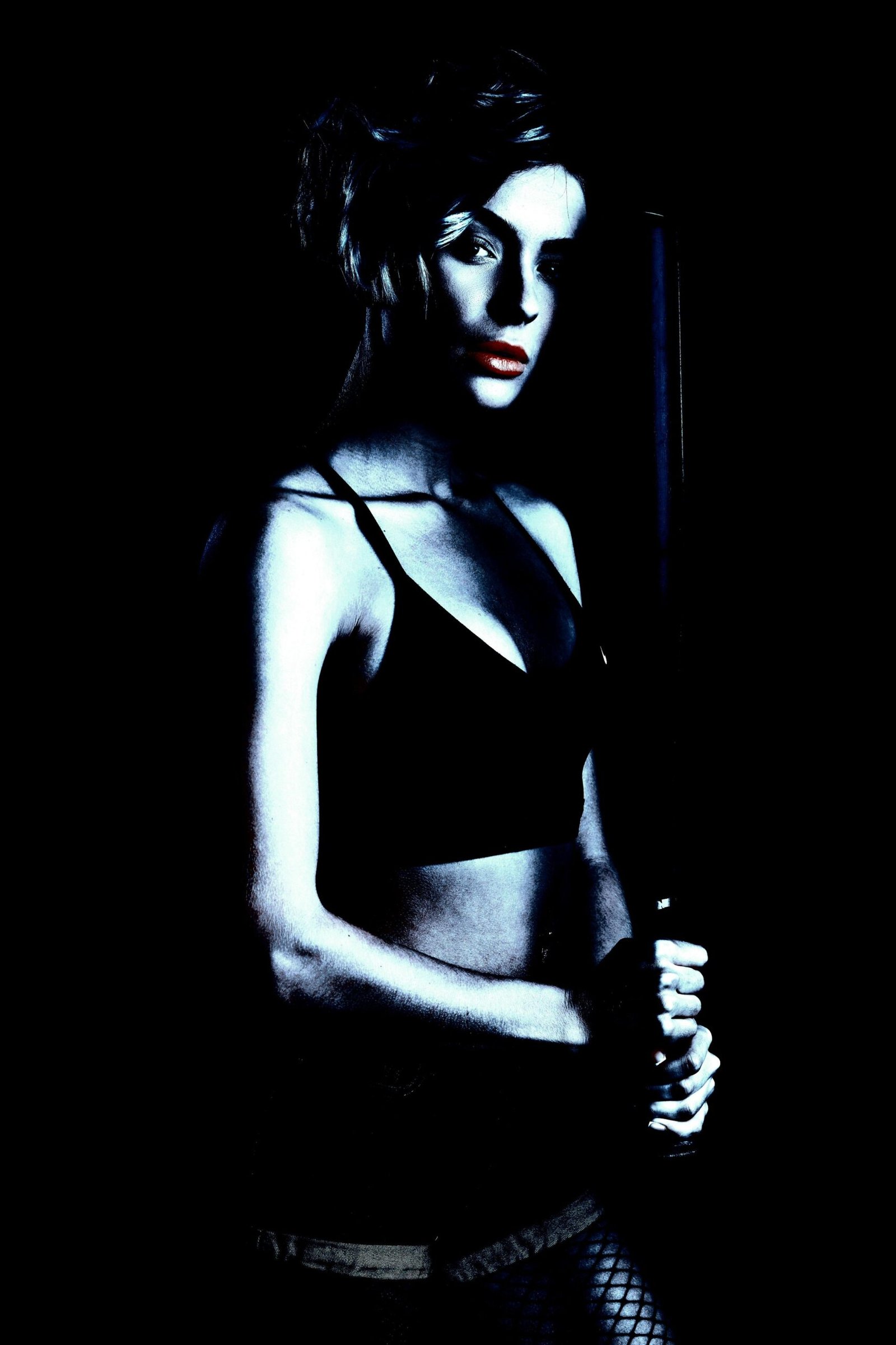 Dark, mysterious woman holding a baseball bat, dressed in athletic wear and fishnet stockings against a black backdrop.