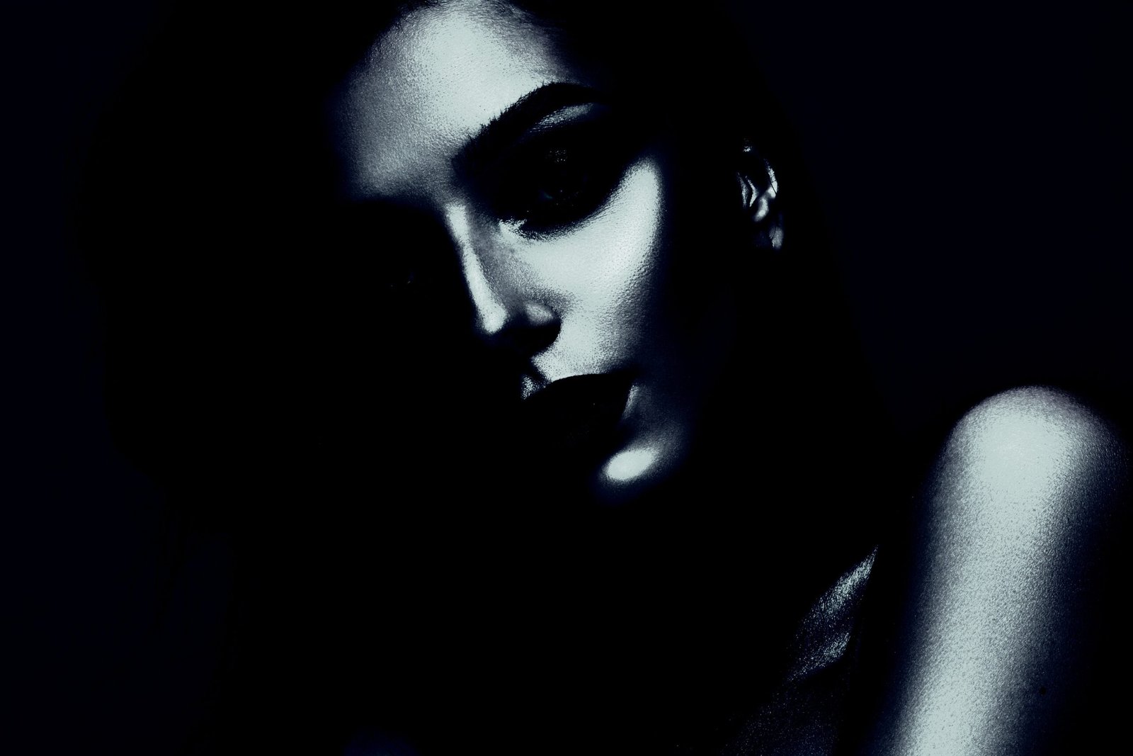Close-up of a woman's face half-hidden in darkness, with a moody, monochromatic blue tone highlighting her features.