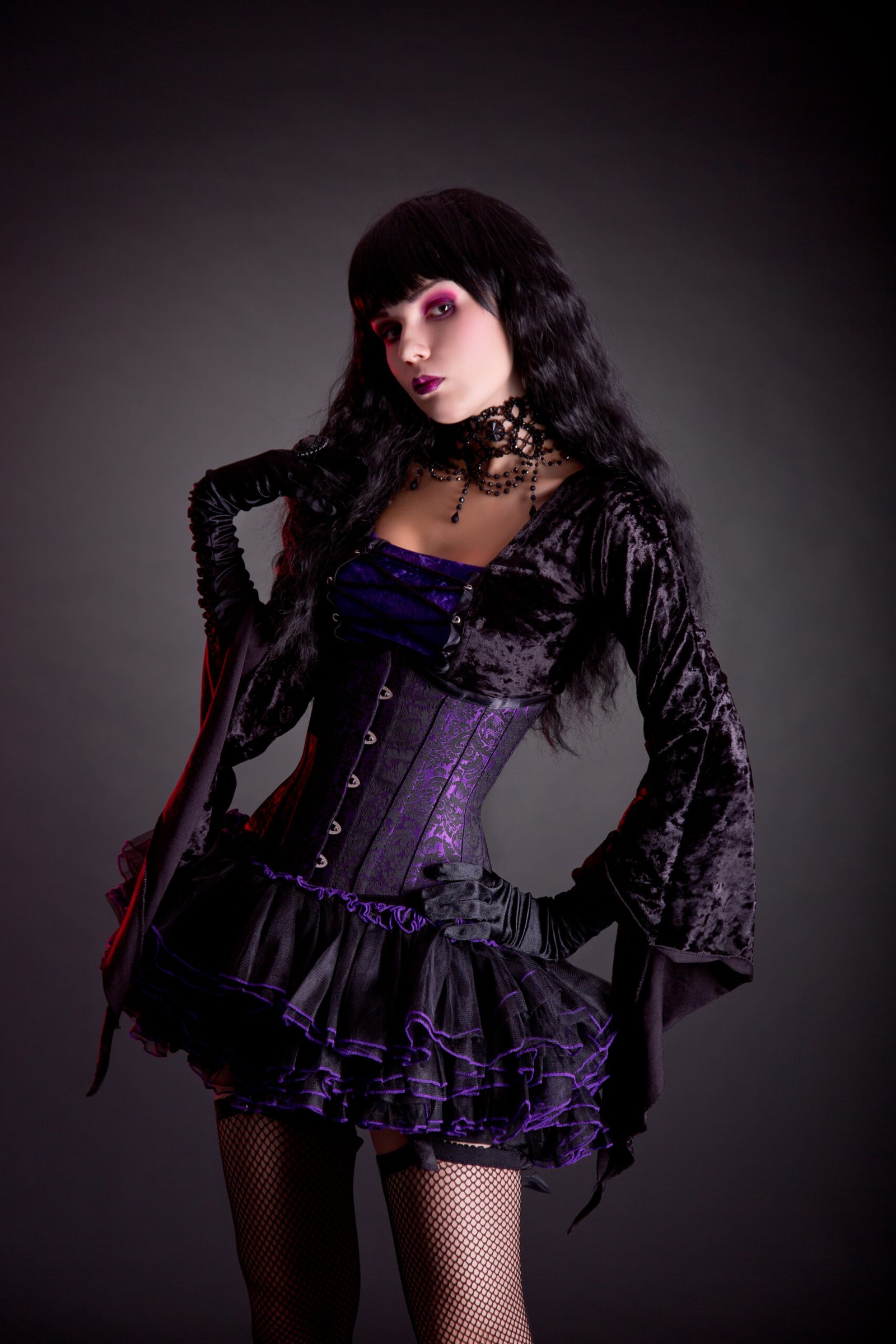 Woman dressed in a gothic outfit with a purple corset and black lace, posing confidently against a dark backdrop.