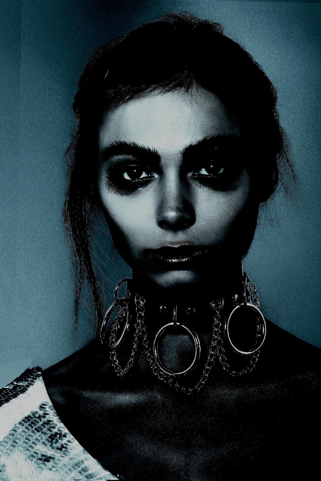 A woman in a high-contrast image wearing a gothic chain choker with large metallic rings, set against a dark blue background.