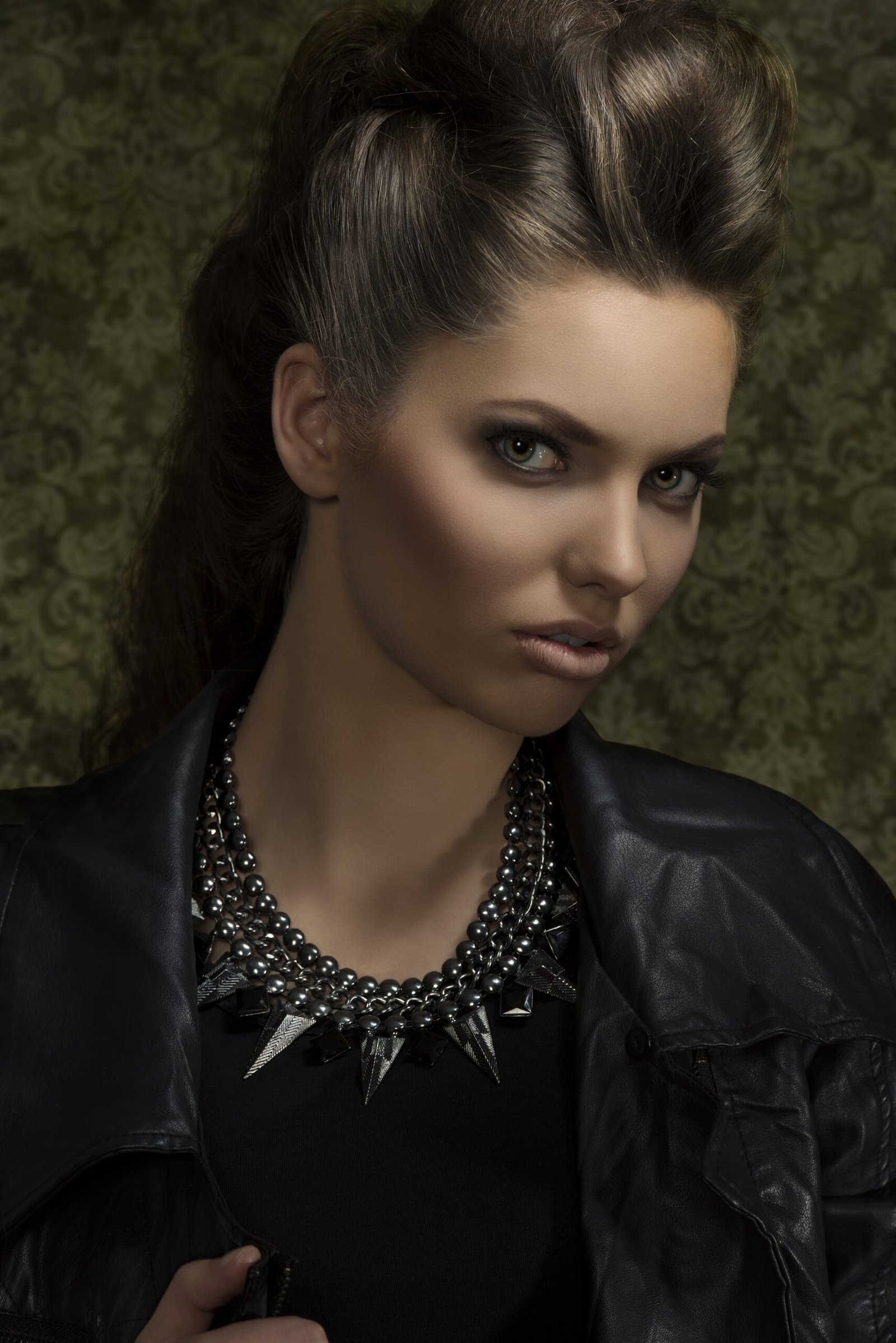 Woman with styled hair and a statement spiked necklace, wearing a leather jacket, exuding a dark, edgy look.