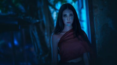 A woman standing in a dimly lit forest, her face calm yet haunting against the blue-toned background, dressed in a draped red cloth.