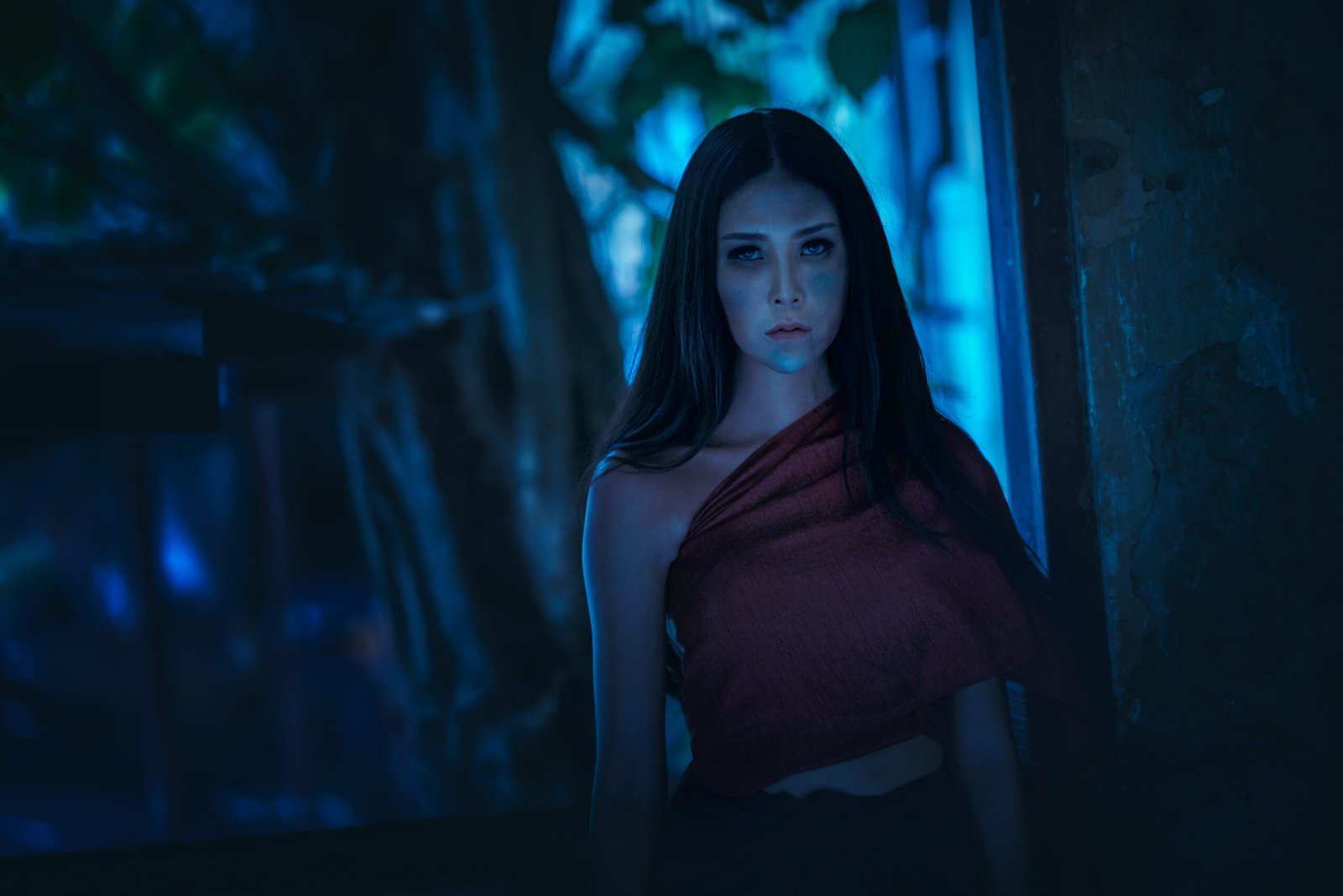 A woman standing in a dimly lit forest, her face calm yet haunting against the blue-toned background, dressed in a draped red cloth.