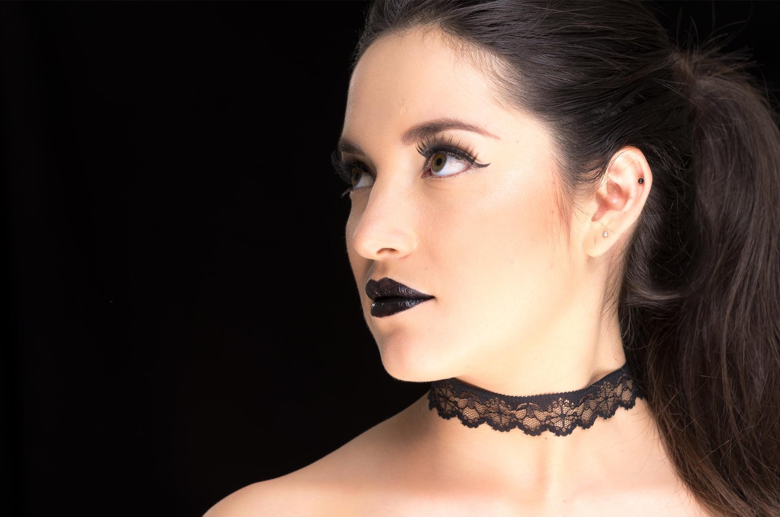 A woman with dark hair wearing a black lace choker, bold black lipstick, and dramatic eyeliner, exuding a classic gothic aesthetic.