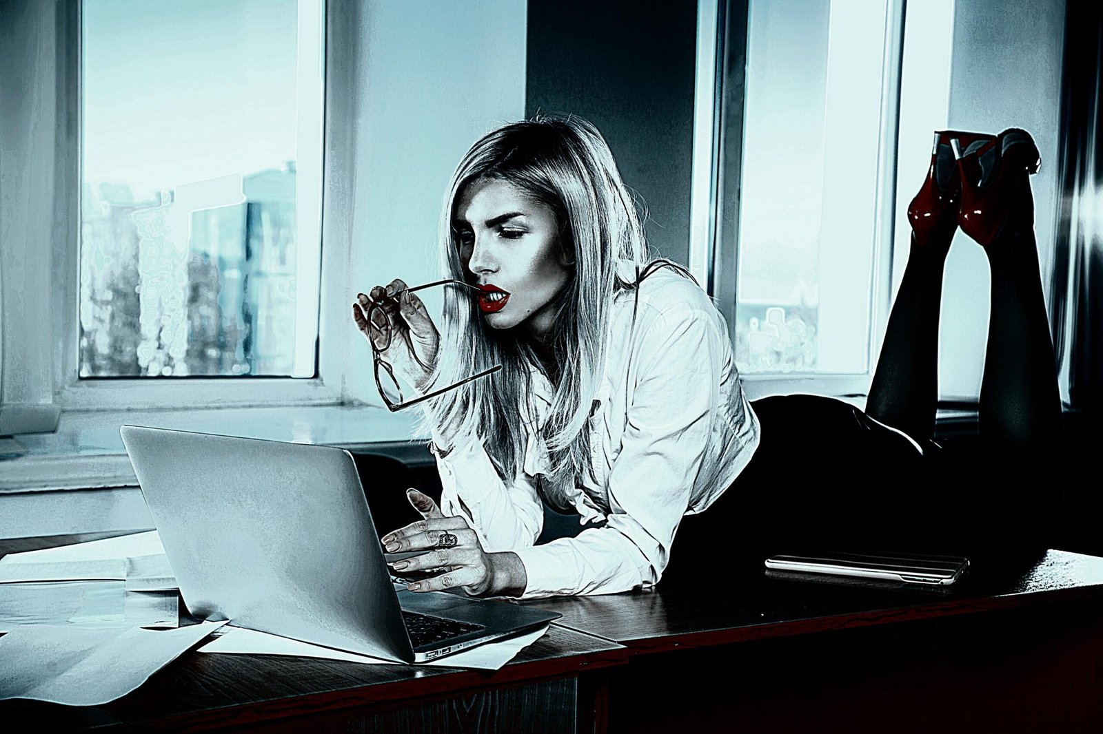 Professional woman lounging on a desk with a laptop, holding glasses and exuding confidence and style.