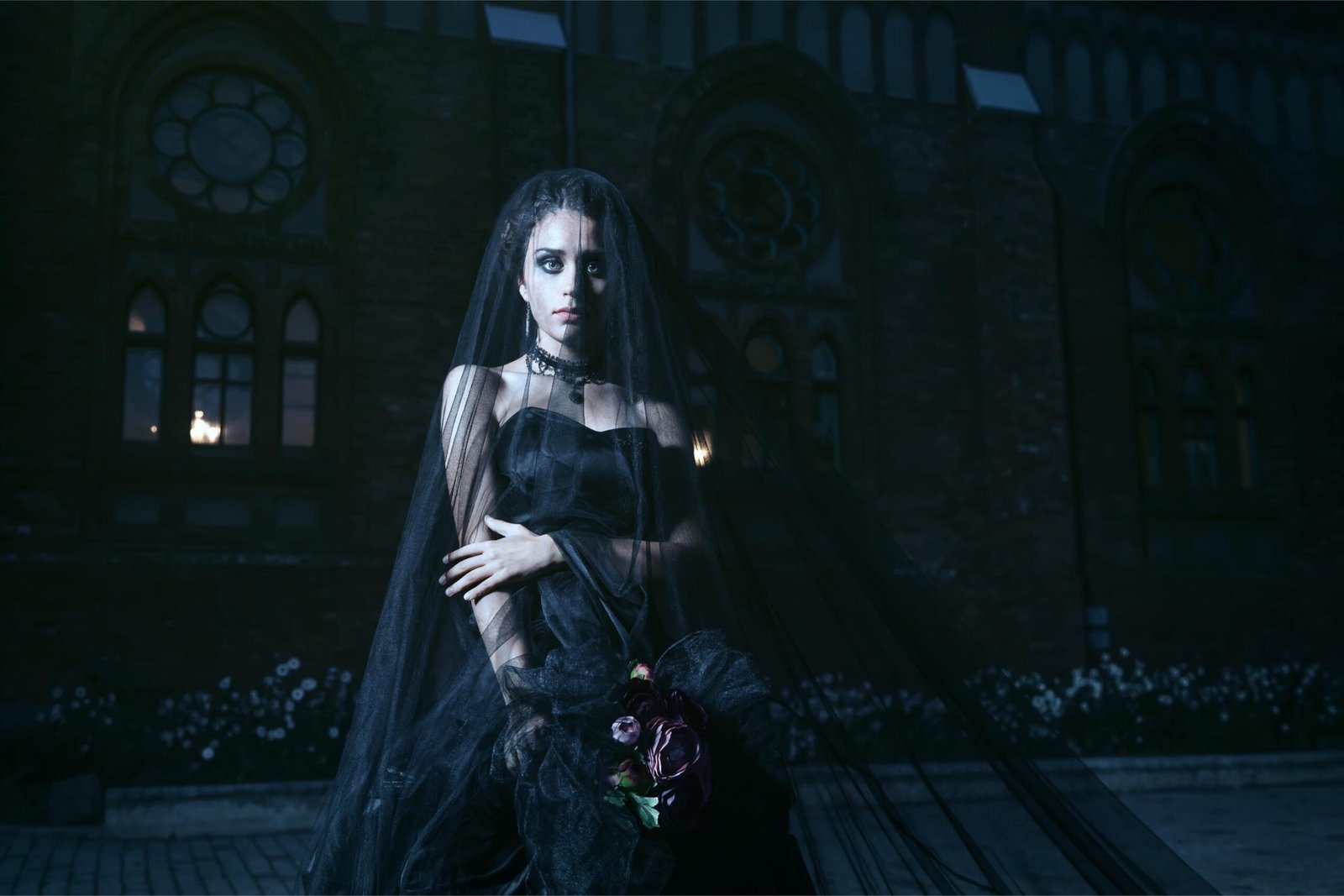A gothic bride dressed in a black veil and flowing dress, standing solemnly in front of an old church at night, holding dark flowers.