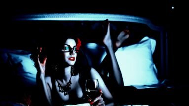 A glamorous woman in glasses and dark attire lounges on a bed, holding a glass of wine, combining elegance with a sultry, enigmatic look.