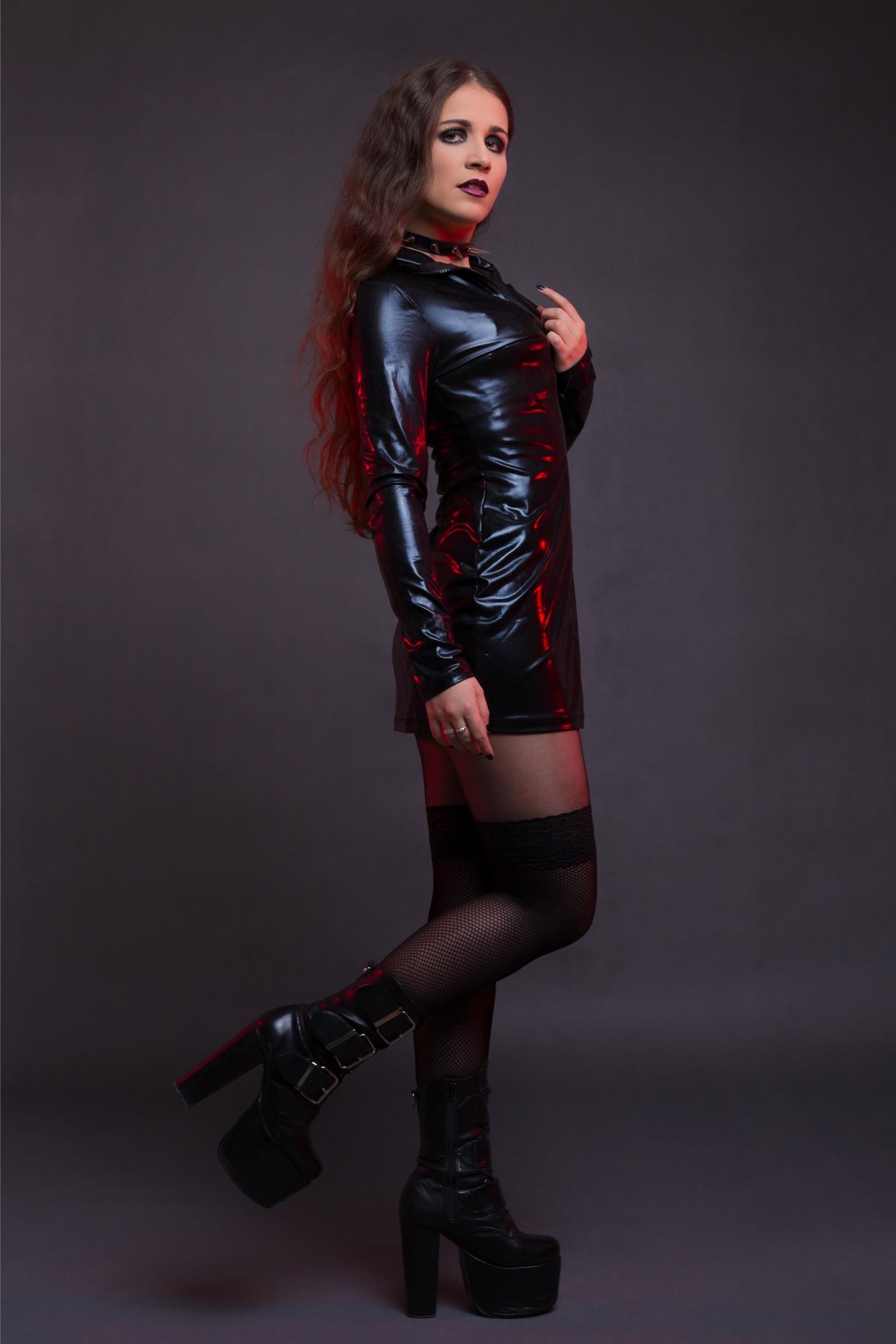 A woman dressed in a sleek, shiny black latex dress with a high neckline, dark makeup, and platform boots, posing confidently against a dark background.