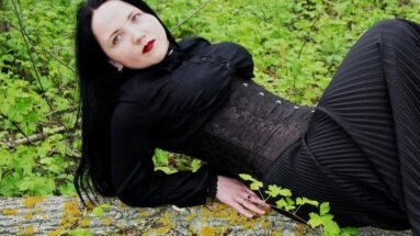 Woman in black underbust corset and dark flowing dress reclining on a mossy tree trunk in a lush green forest.