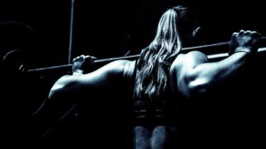 Back view of a woman lifting a heavy barbell in a gym, her muscular arms emphasized by dramatic lighting.