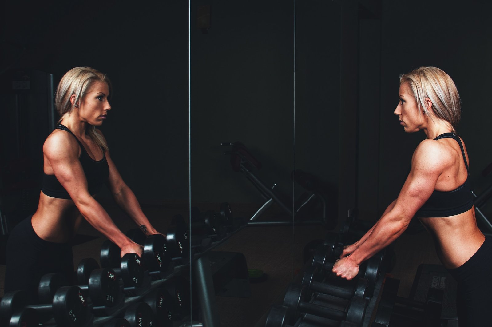 A strong woman leaning on a row of dumbbells in front of a mirror, wearing black athletic wear, focused on her workout.