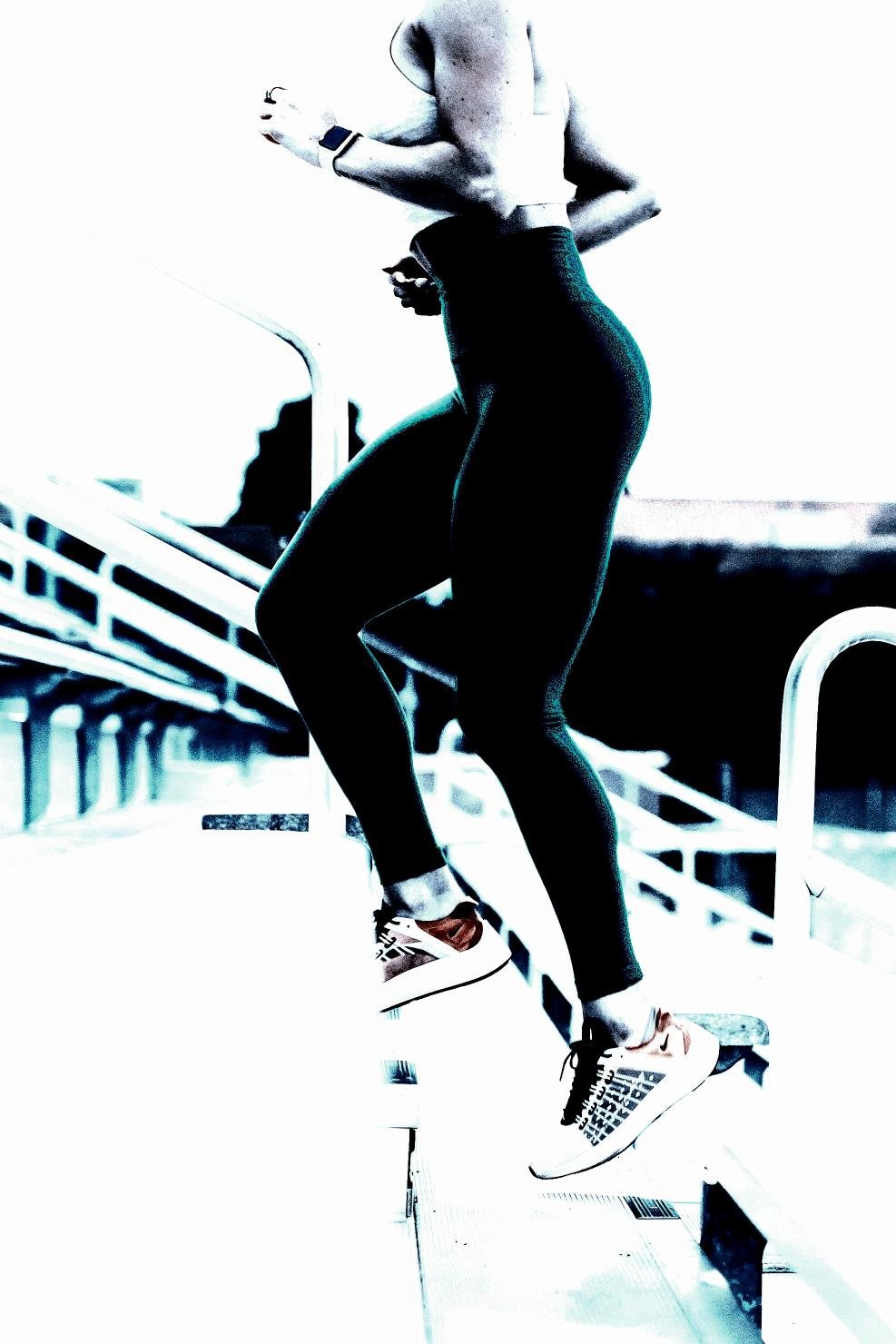 Woman running up stairs outdoors, wearing sleek athletic leggings and trainers, framed by soft lighting.