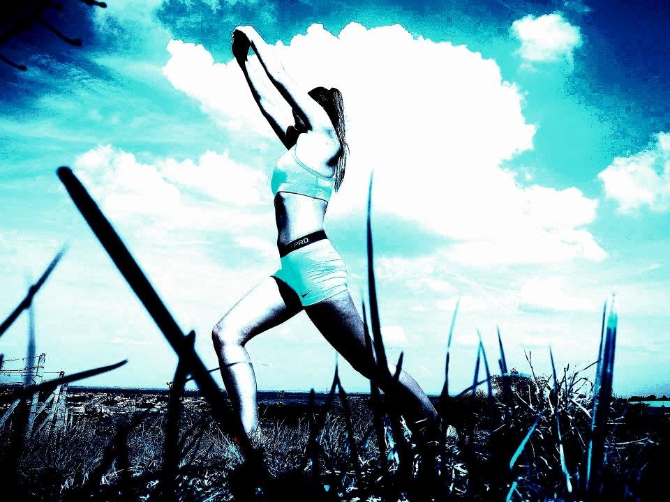 Woman stretching in an open field, wearing light workout gear, with dramatic contrasts and a cloudy sky in the background.