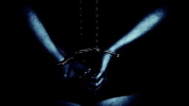 A person in a corset with hands cuffed behind their back, captured in moody, high-contrast lighting.