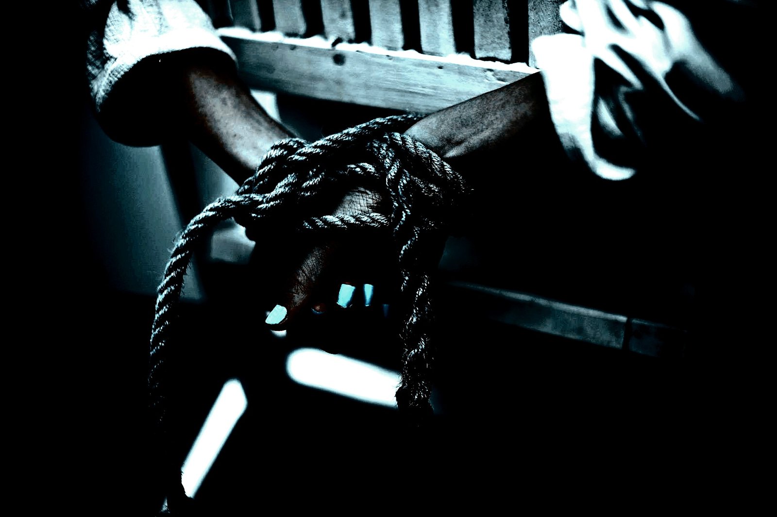 Hands bound with thick rope against a wooden bedframe, emphasizing tension and submission.