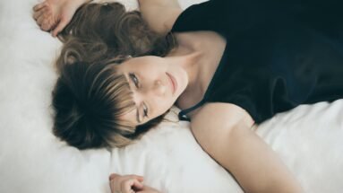 Woman in a black satin nightgown lying on a white bed, gazing softly at the camera with her hair spread out.