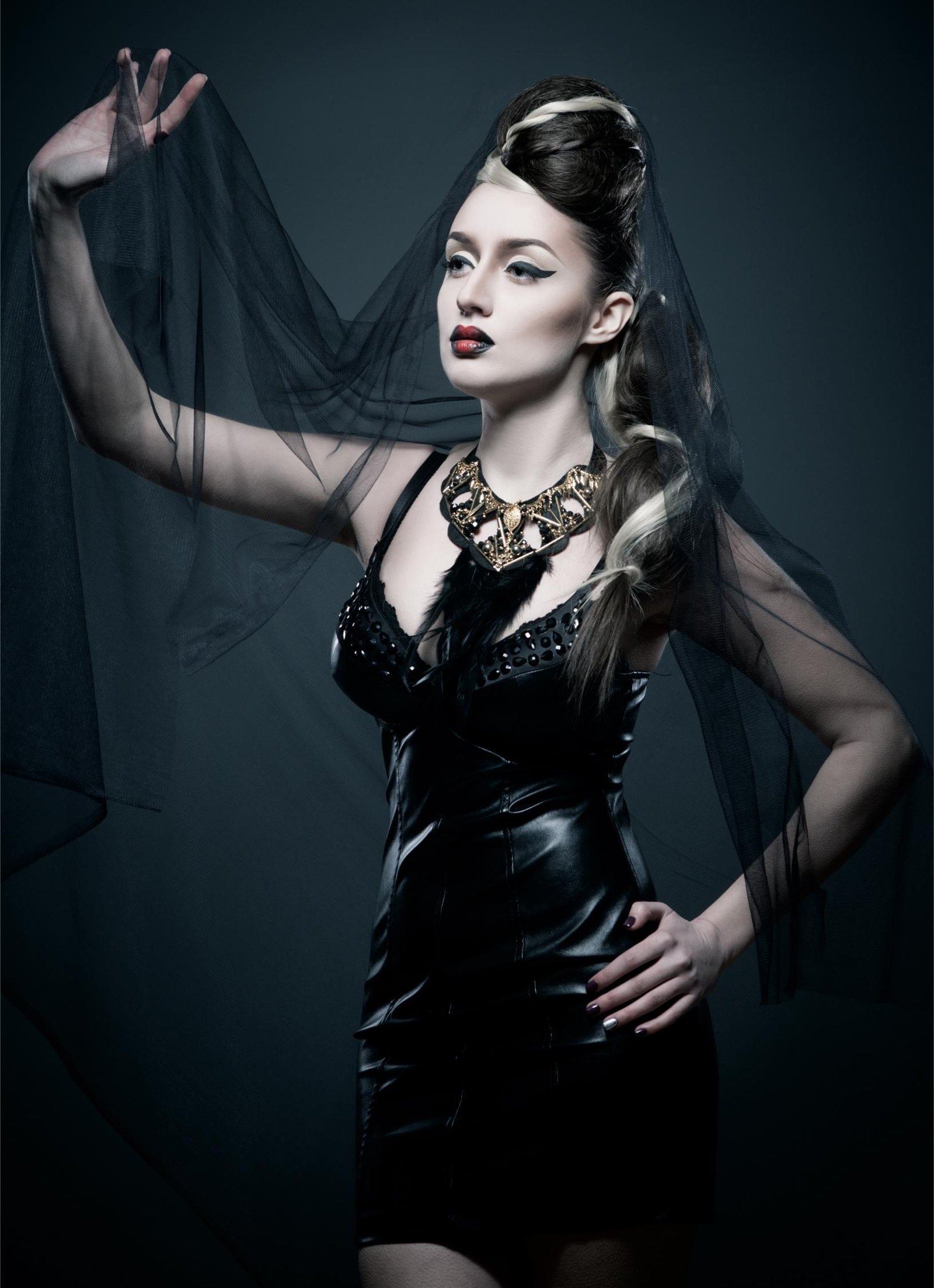Woman in a gothic veil and a black dress with a statement necklace, posing with an intense expression and red lipstick.
