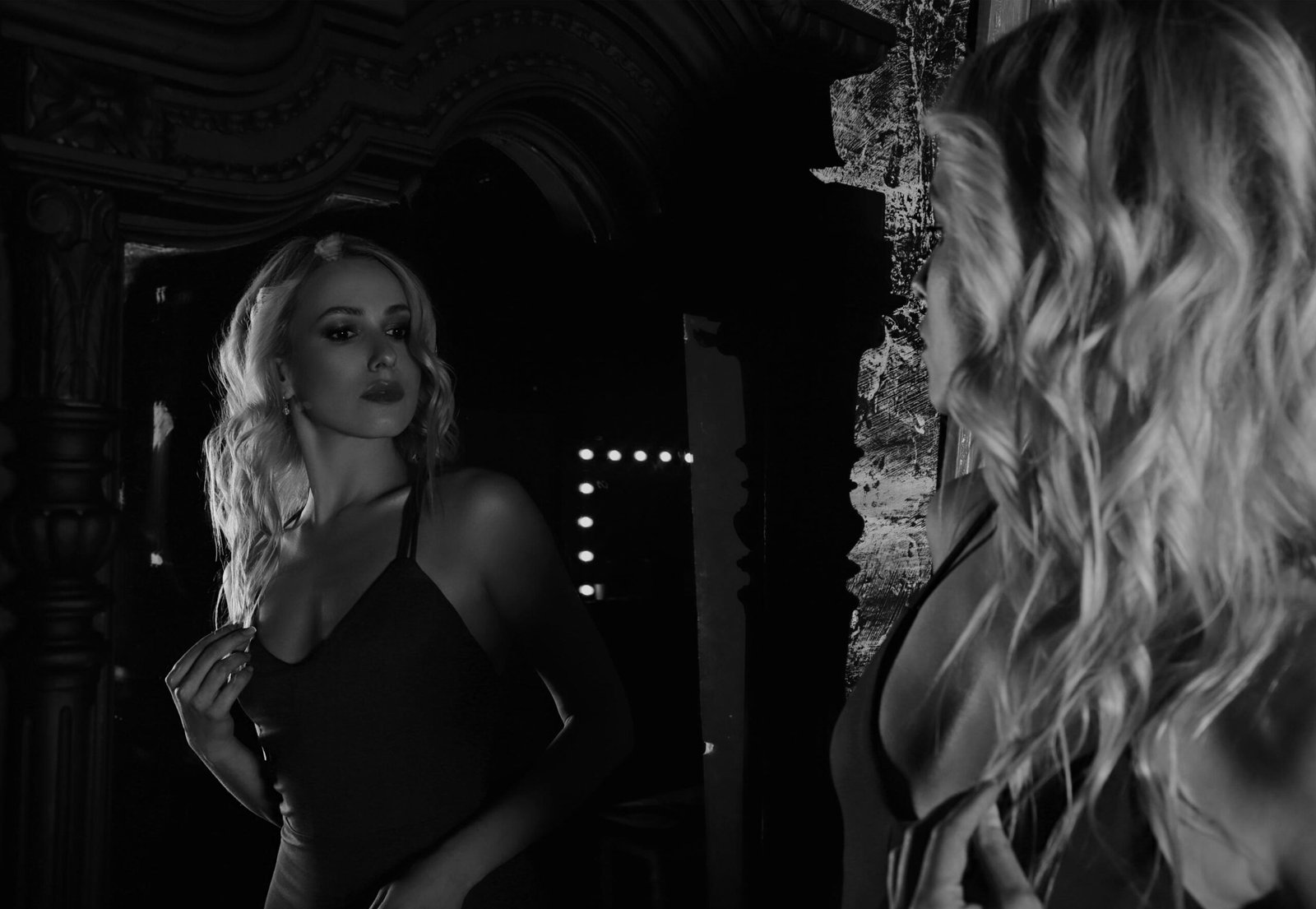 Woman in a dark dress with wavy blonde hair, gazing at her reflection in a vintage mirror in a dimly lit room.