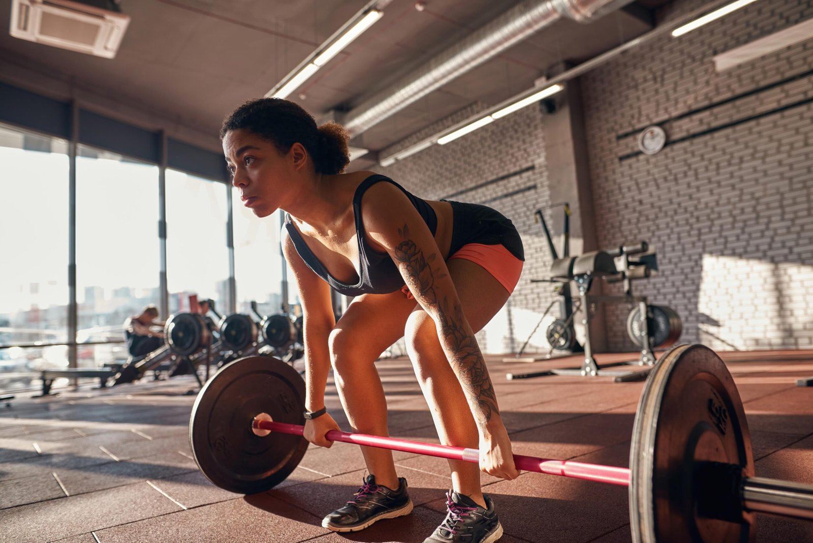 Focused woman with tattoos lifting a barbell in a gym, wearing a black tank top and pink shorts, with intense natural lighting.