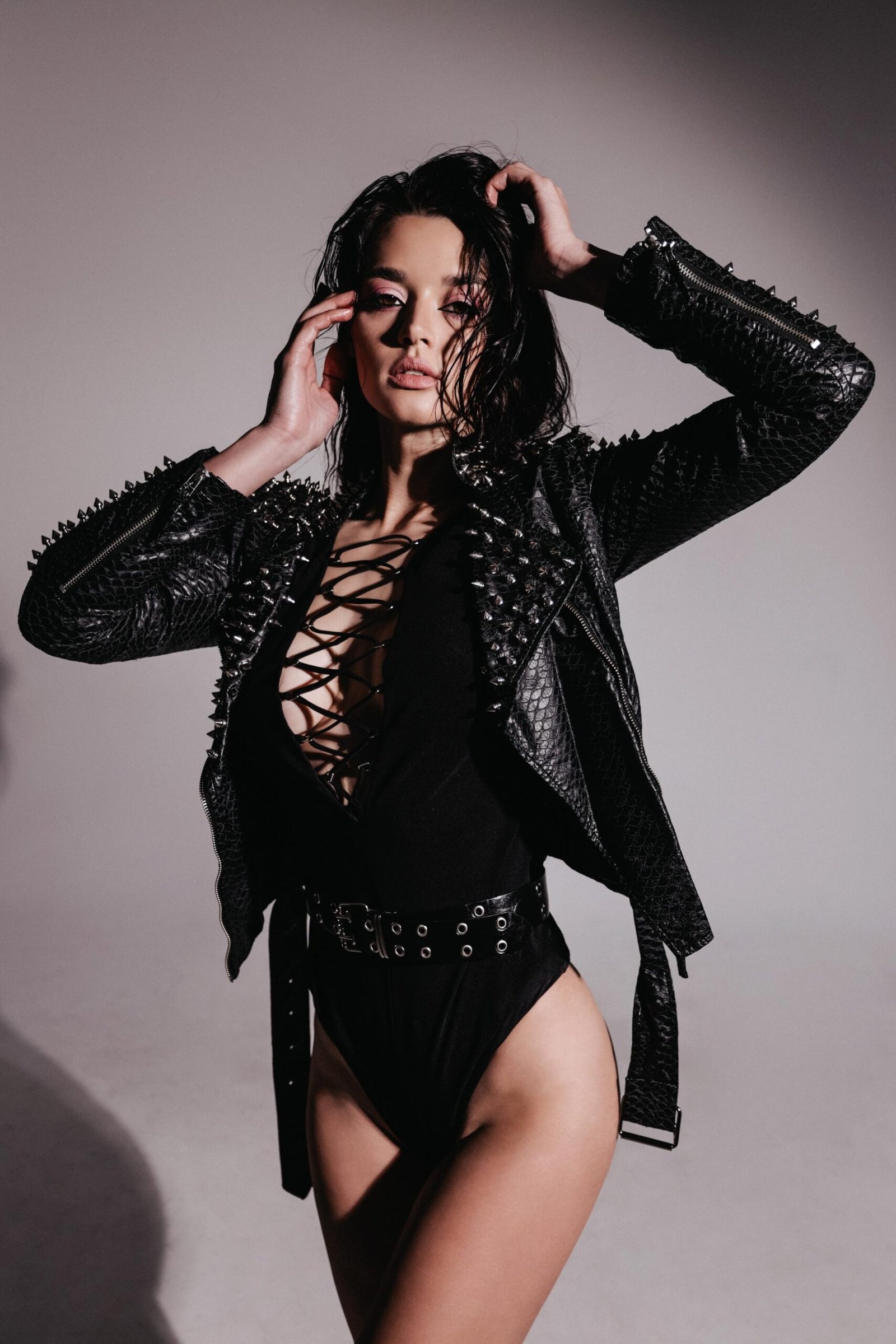 Woman in a studded black leather jacket and a laced bodysuit posing confidently, showcasing edgy goth fashion.