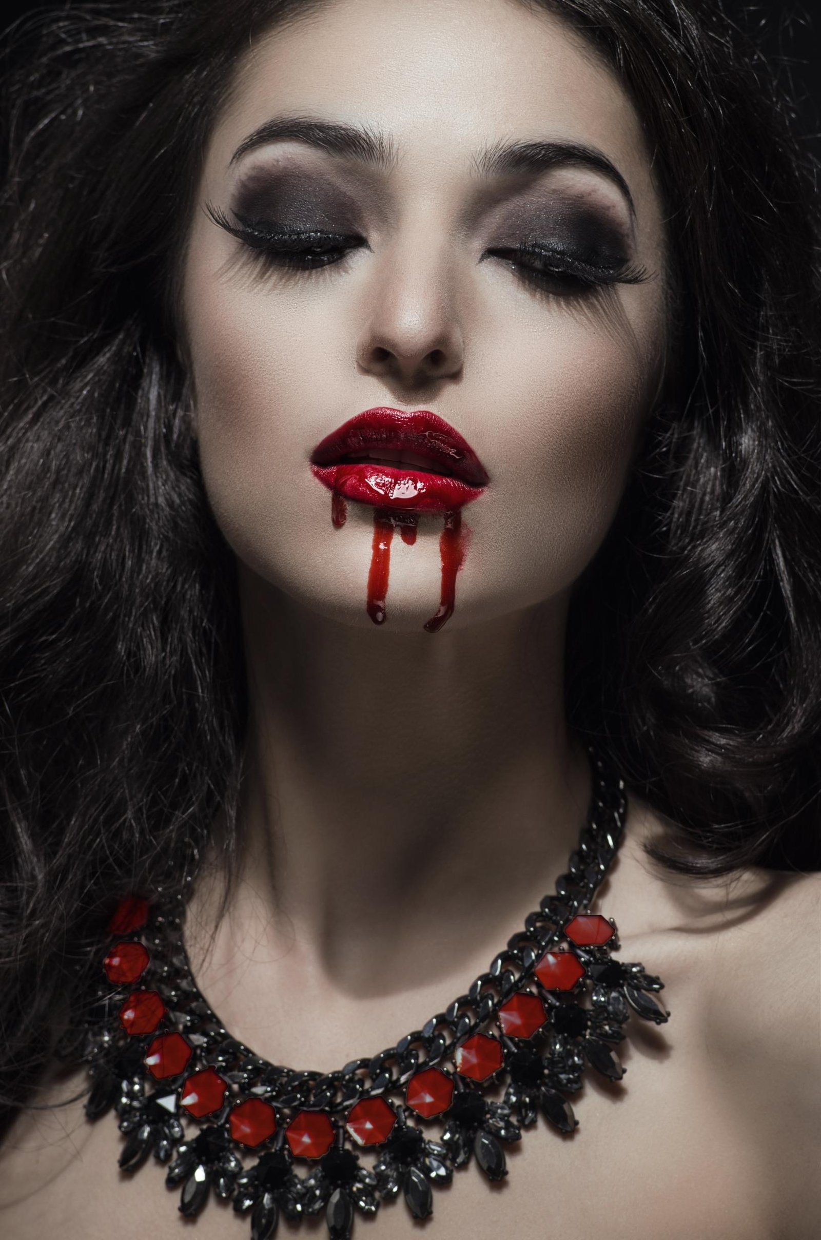 Close-up of a woman with dark makeup and red lips, with blood-like droplets dripping down her mouth.