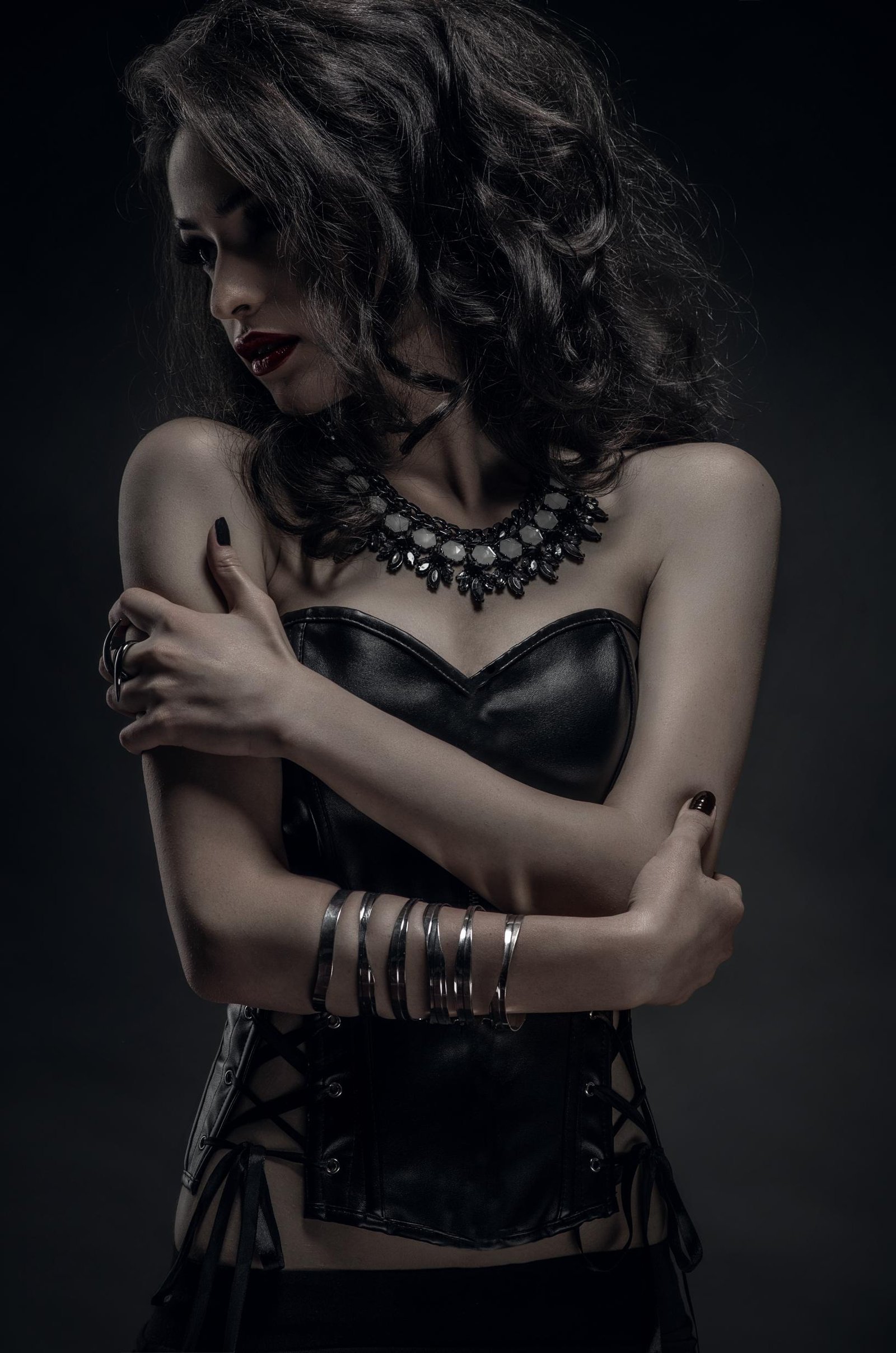 Woman in a corset with arms crossed, wearing dark makeup and a black necklace, gazing downward in a dramatic pose.