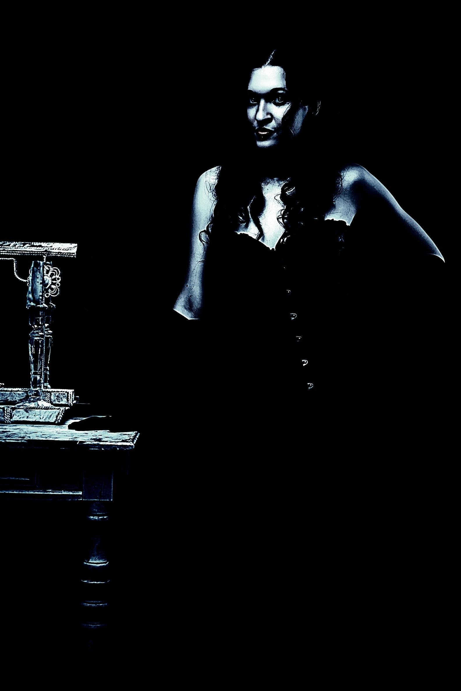 Gothic woman in a corset dress standing near a vintage table, shrouded in shadow with a moody and eerie aesthetic.