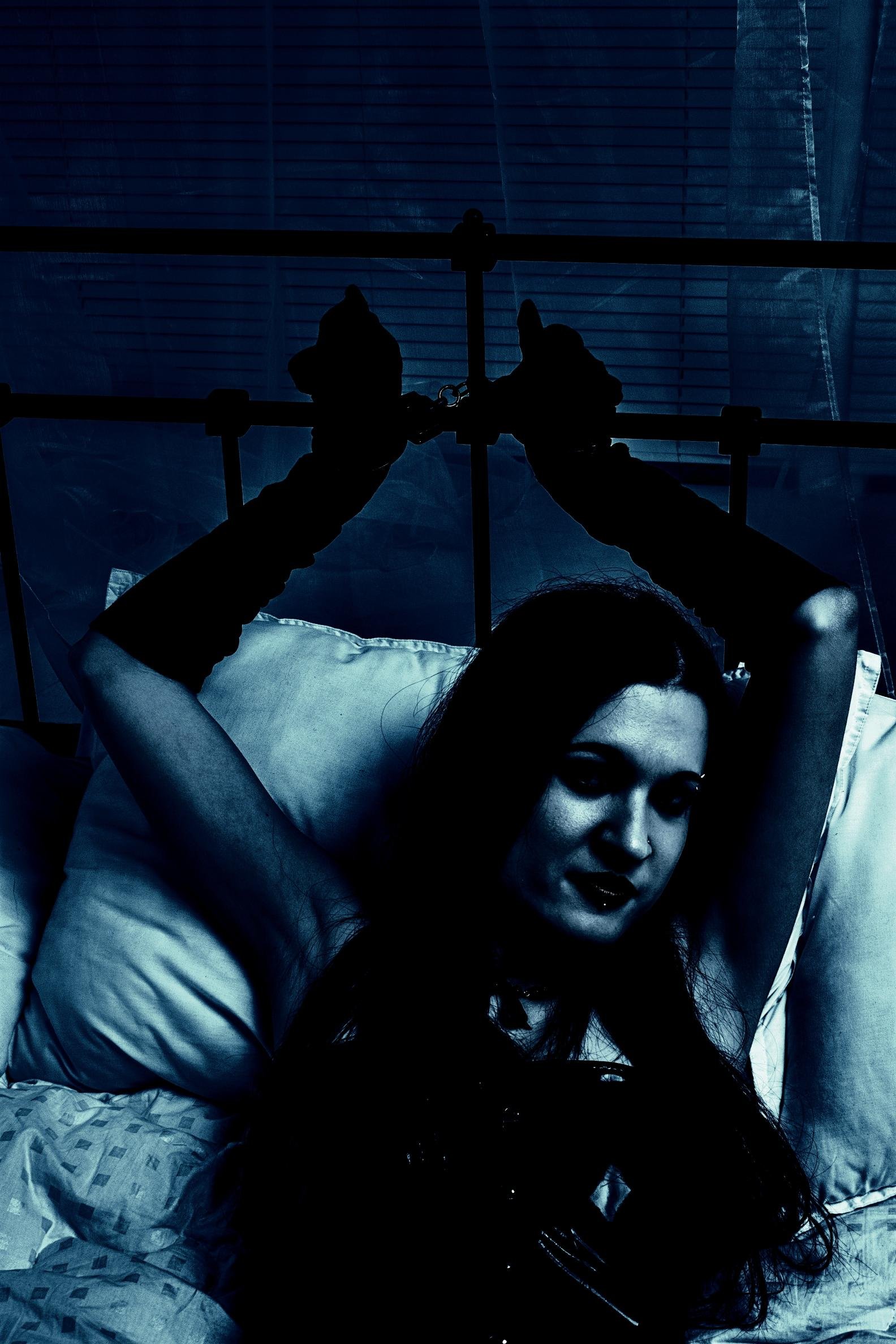 Dark-toned silhouette of a woman in handcuffs, lying on a bed with a mysterious, gothic atmosphere.