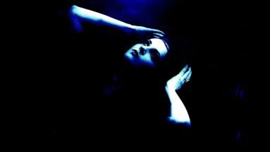 Woman lying back with hands in hair, looking upward in dramatic blue light with a dark, mysterious expression.