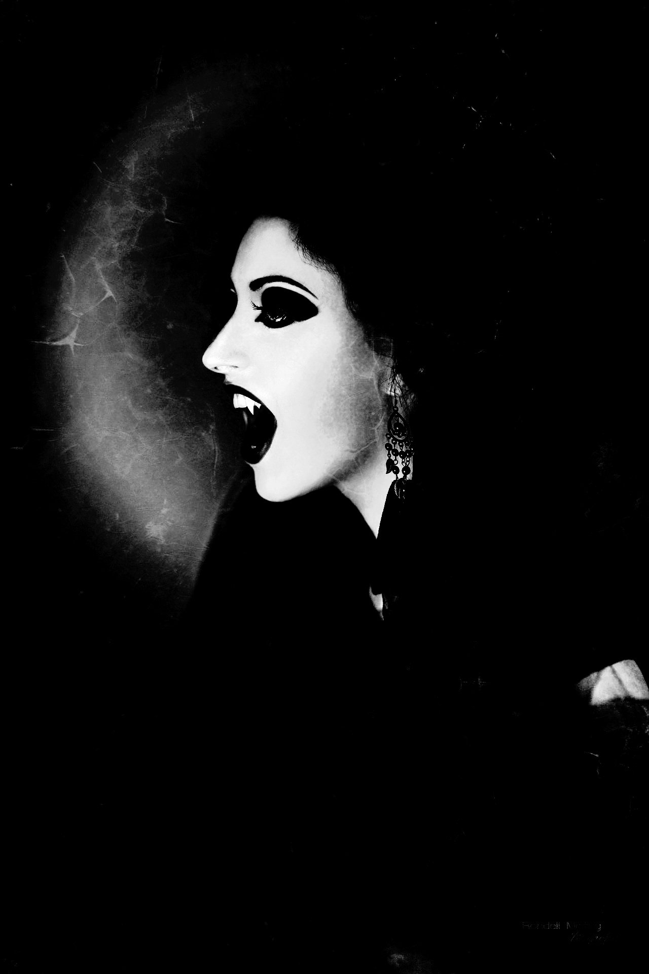 Vampire woman baring fangs in profile with dramatic dark makeup and earrings, set against a shadowy background.