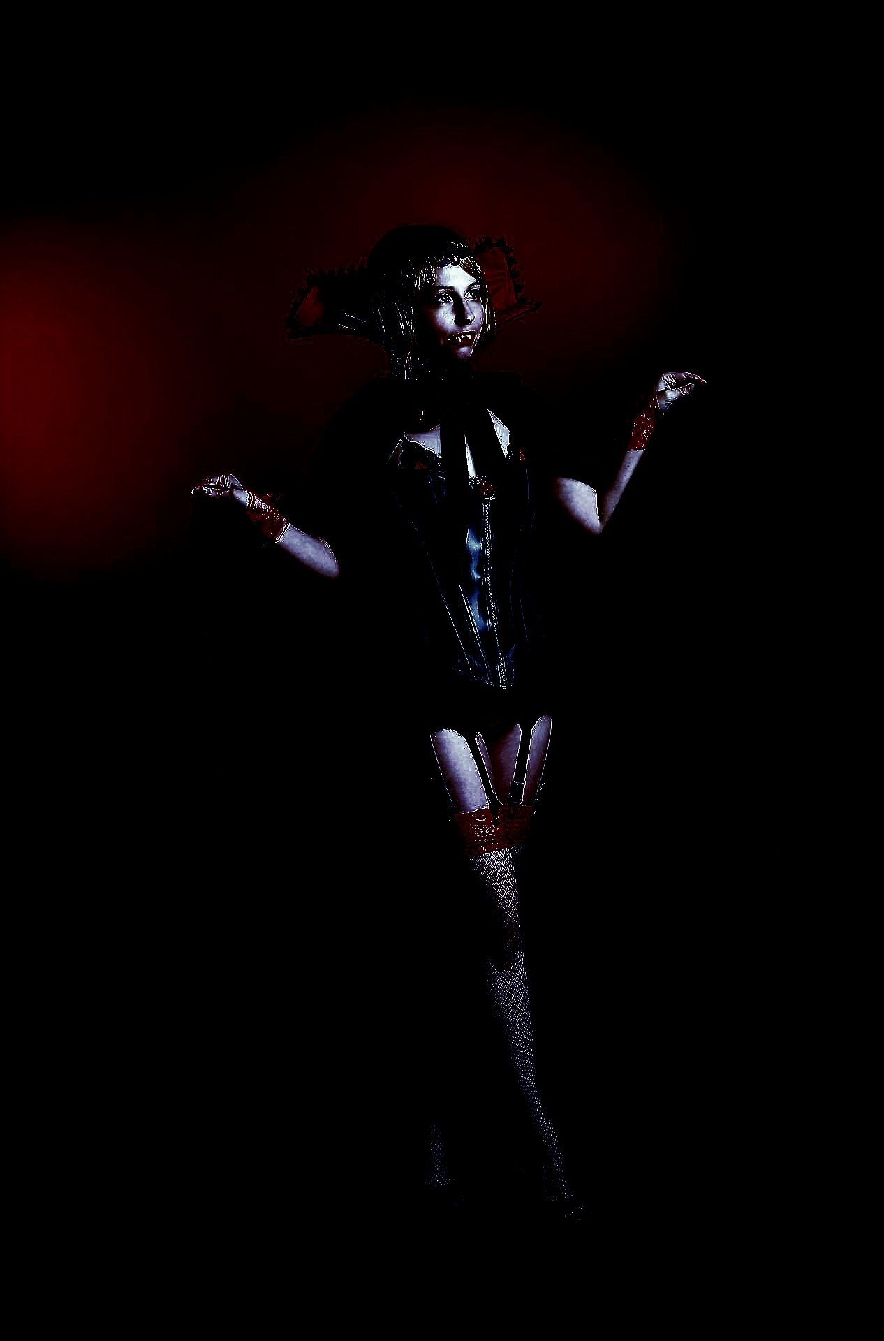 Woman dressed in gothic vampire attire with a red and black theme, posed with arms raised against a dark backdrop.