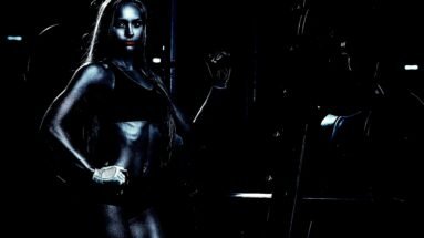 Strong woman in a black sports outfit posing in a dimly lit gym, exuding power and gothic energy.