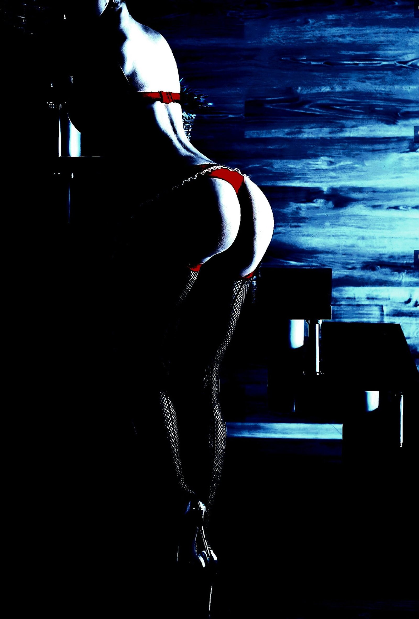 Woman in red lingerie and fishnet tights, leaning on a staircase in a moody, low-light setting.